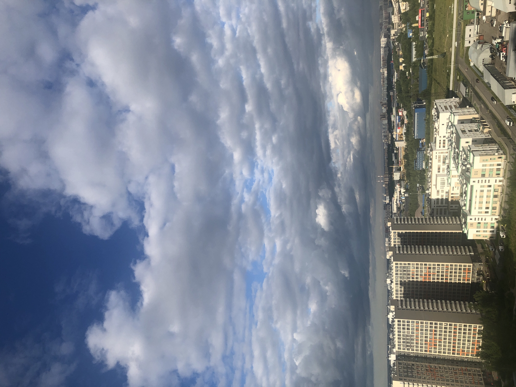 The image shows an aerial view of a city with a dramatic sky. The sky is filled with a mix of dark, heavy clouds and bright, white cumulus clouds, suggesting a dynamic weather system. Sunlight breaks through in places, casting light and shadow on the landscape below.
The city appears to be a mix of residential and commercial buildings. Tall, modern apartment buildings with balconies and many windows are prominent, some with light-colored facades and others darker. There are also lower-rise buildings, some with industrial or commercial appearances, and green spaces with trees interspersed throughout. Roads with cars are visible, indicating ongoing activity.
The orientation of the image is vertical, as if taken from a very high vantage point looking down and to the side. The time of day is likely afternoon or late afternoon, given the lighting conditions and cloud formations. The weather appears changeable, with potential for rain or storms due to the dark clouds, contrasted with bright sunlight. No people are clearly visible as distinct subjects, but the presence of cars and buildings implies human presence and activity. There is no visible text within the image.