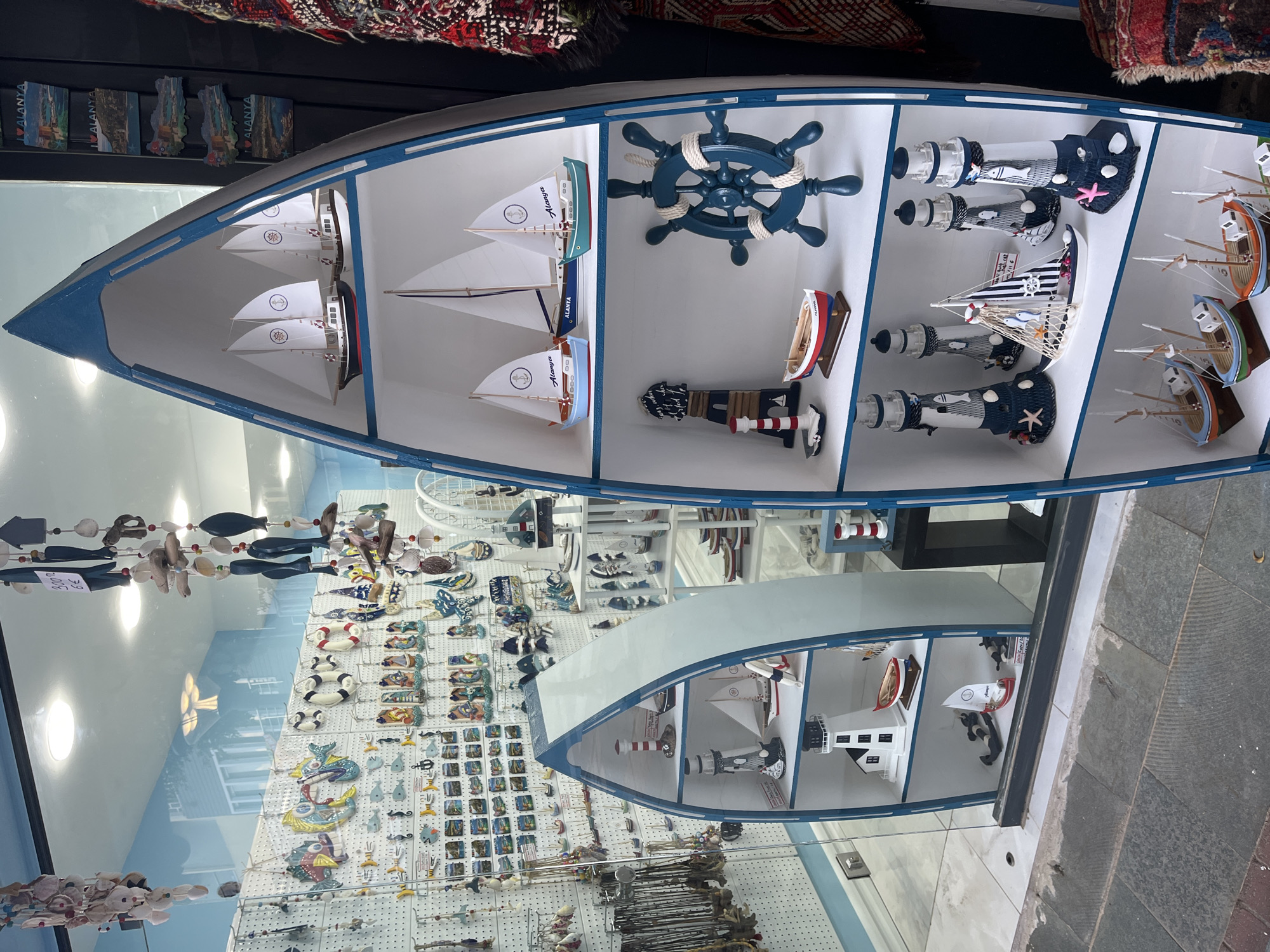 The image shows a storefront display in Alanya, Turkey, featuring a variety of nautical-themed souvenirs. Two boat-shaped shelves dominate the scene. The upper shelf contains model sailboats, a ship's wheel, lighthouses, and model fishing boats. The lower shelf also has model sailboats and lighthouses. Various other souvenirs are visible on the wall behind the boat shelves, including hanging decorations with marine life. The display is set against a backdrop that reflects the street outside.