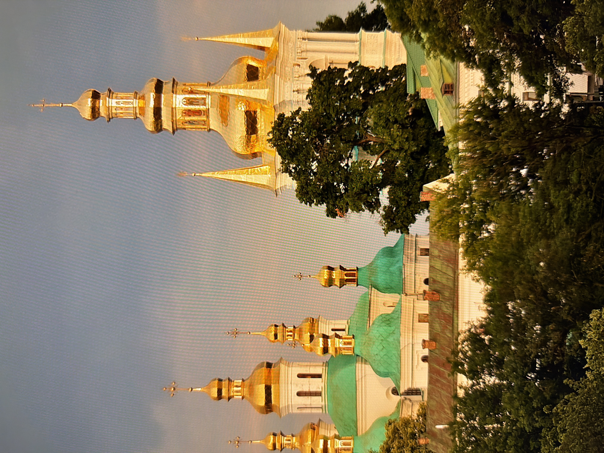 The image depicts the Kyiv Pechersk Lavra, a historic Orthodox Christian monastery located in Kyiv, Ukraine. The scene is dominated by the monastery's iconic golden domes and white structures, set against a hazy blue sky. Several green-roofed buildings are also visible, characteristic of Ukrainian architecture. Lush green trees partially obscure the lower levels of the monastery, suggesting a viewpoint from a slightly elevated position or surrounded by greenery. The lighting suggests it might be late afternoon or early morning, with a soft, warm glow illuminating the golden domes. No people are visible in the frame. The overall atmosphere is serene and majestic, highlighting the architectural grandeur of the monastery.