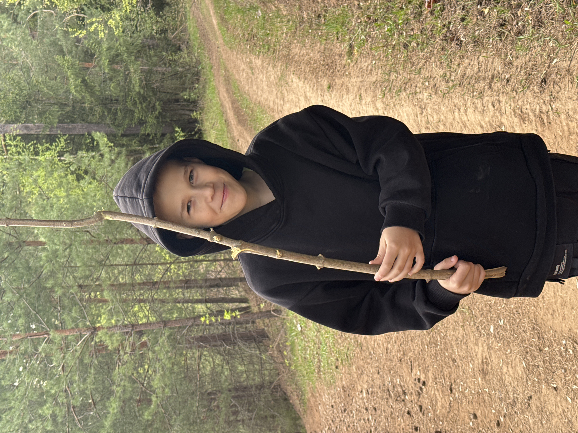 A young child stands on a dirt path in a wooded area, holding a stick horizontally. The child is wearing a black hooded sweatshirt and dark pants. The path runs through a forest of pine trees with sparse green foliage. The ground is covered in dry grass and fallen leaves. This photo was likely taken in Kamennoye, Russia.