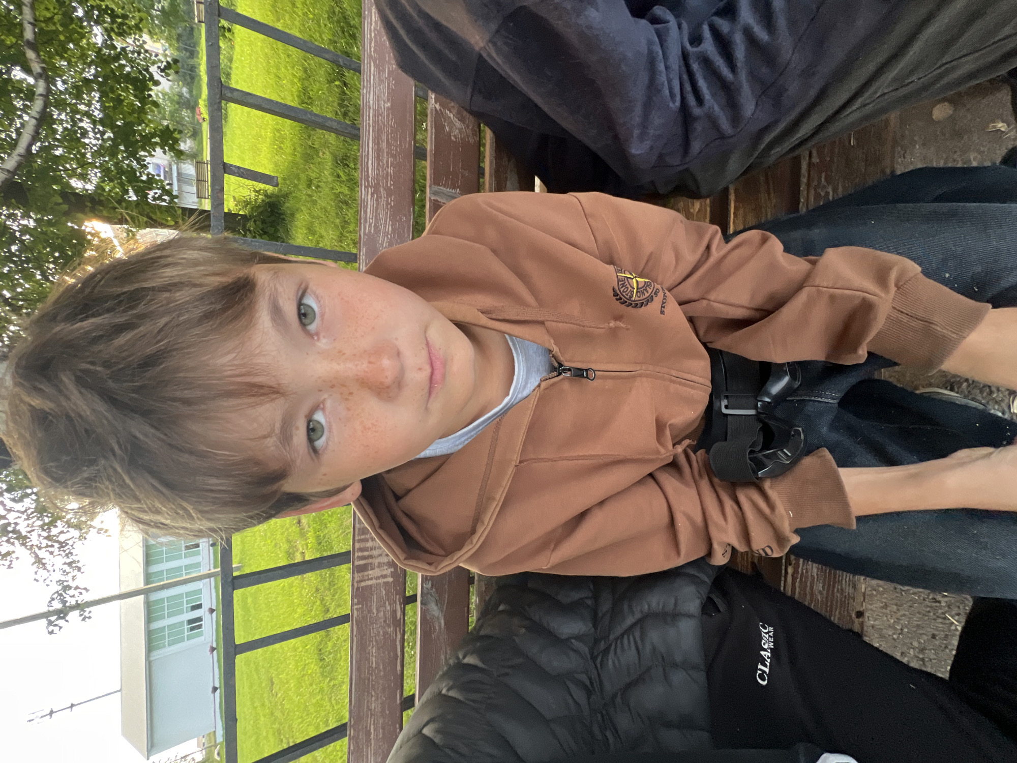The media file shows a close-up shot of a young boy sitting on a wooden bench outdoors. He has light brown hair with bangs, green eyes, and freckles across his nose and cheeks. He is wearing a brown hooded sweatshirt over a white t-shirt and dark blue jeans. The sweatshirt has a circular logo on the left chest. A black belt is visible around his waist.
The background features a grassy area with trees and a building with large windows, suggesting a park or outdoor recreational area. The lighting indicates it might be late afternoon or early evening, with the sun casting a warm glow. The overall mood appears casual and relaxed.
The image was taken in Perevoznoe, Russia. There is text visible on the black puffer jacket in the foreground which reads "CLASSIC WEAR".