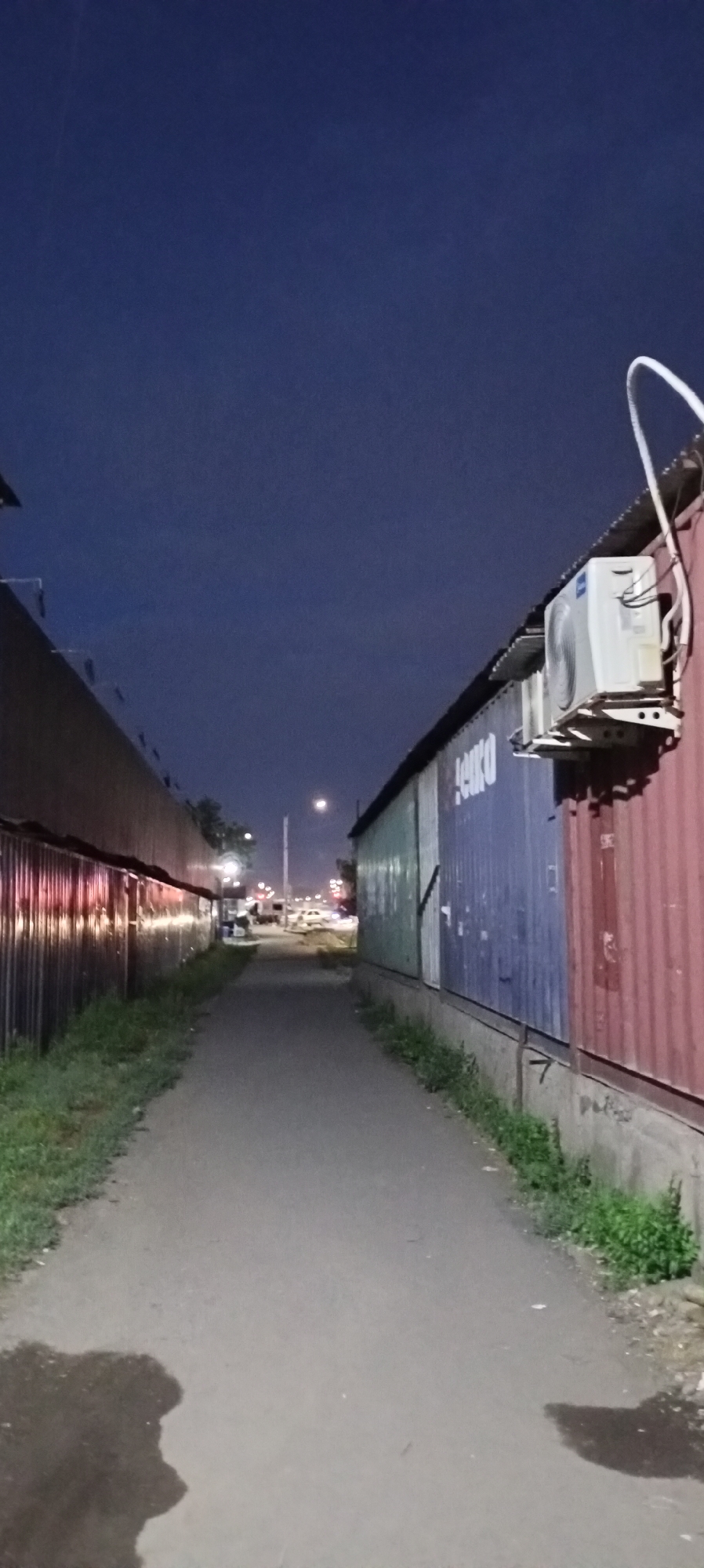 The image shows a narrow, unpaved path between two corrugated metal structures at dusk. The structure on the left is a dark, metallic wall with a reflection of red and orange lights. The structure on the right is composed of shipping containers, one green, one blue with the word "EURO" visible, and one red. An air conditioning unit is mounted on the side of the red shipping container. The sky is a deep, twilight blue. In the distance, there is a street with vehicles and a lamppost, suggesting a roadside location. The path itself has some scattered debris and weeds growing along the edges, particularly in front of the shipping containers. A shadow of what appears to be a person is visible on the lower left portion of the path. The scene is characteristic of an industrial or commercial area on the outskirts of a city, possibly near a port or logistics hub. This location is identified as being in Sokuluk, Kyrgyzstan.