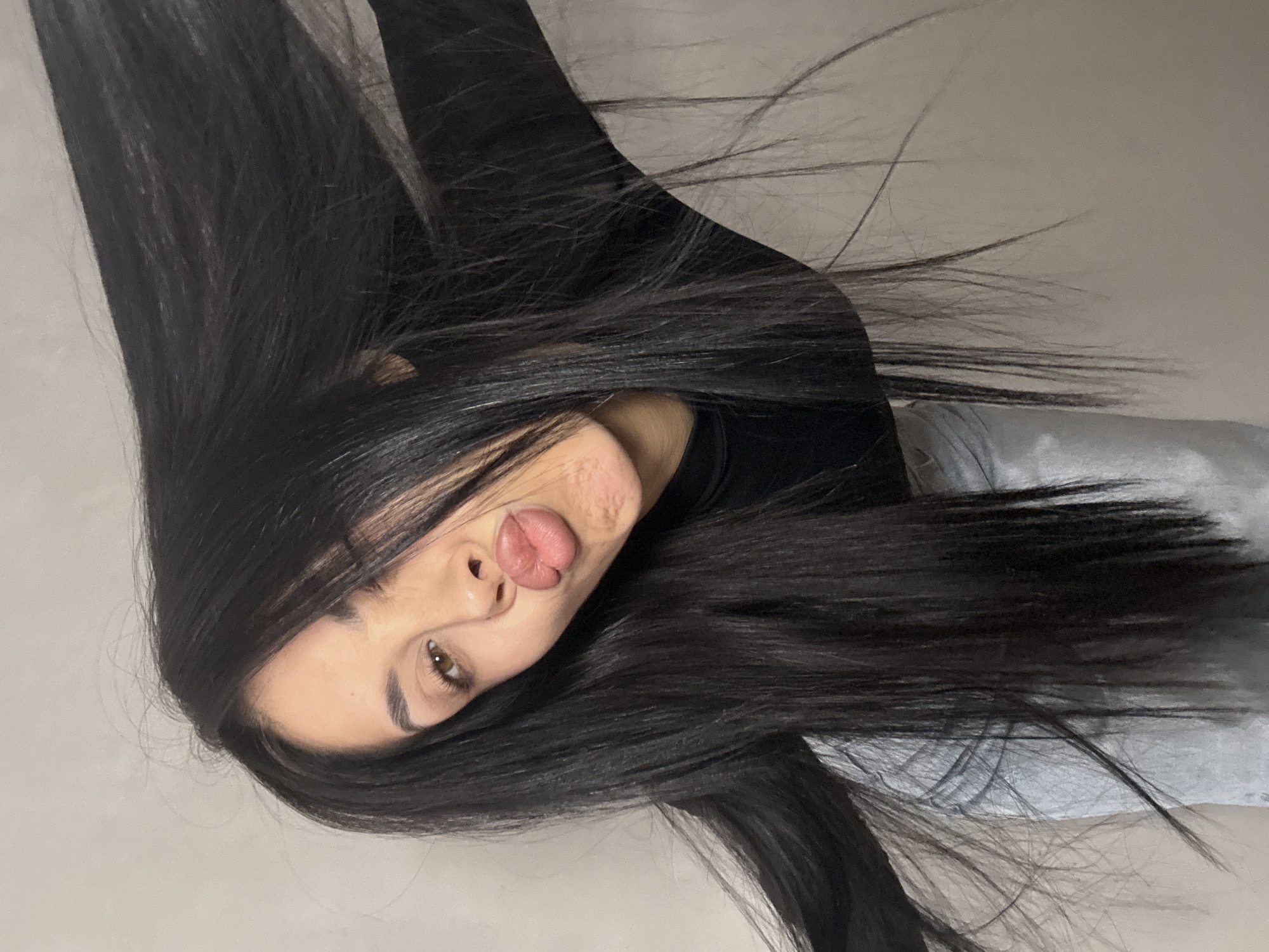 A young woman with long, dark hair, is posing with her lips pursed in a kiss. Her right eye is partially closed, and her hair is being thrown back, obscuring part of her face. She is wearing a black top and light-colored jeans. The background is a plain, light-colored wall. There is no indication of the city of Votkinsk, Russia, in this image.