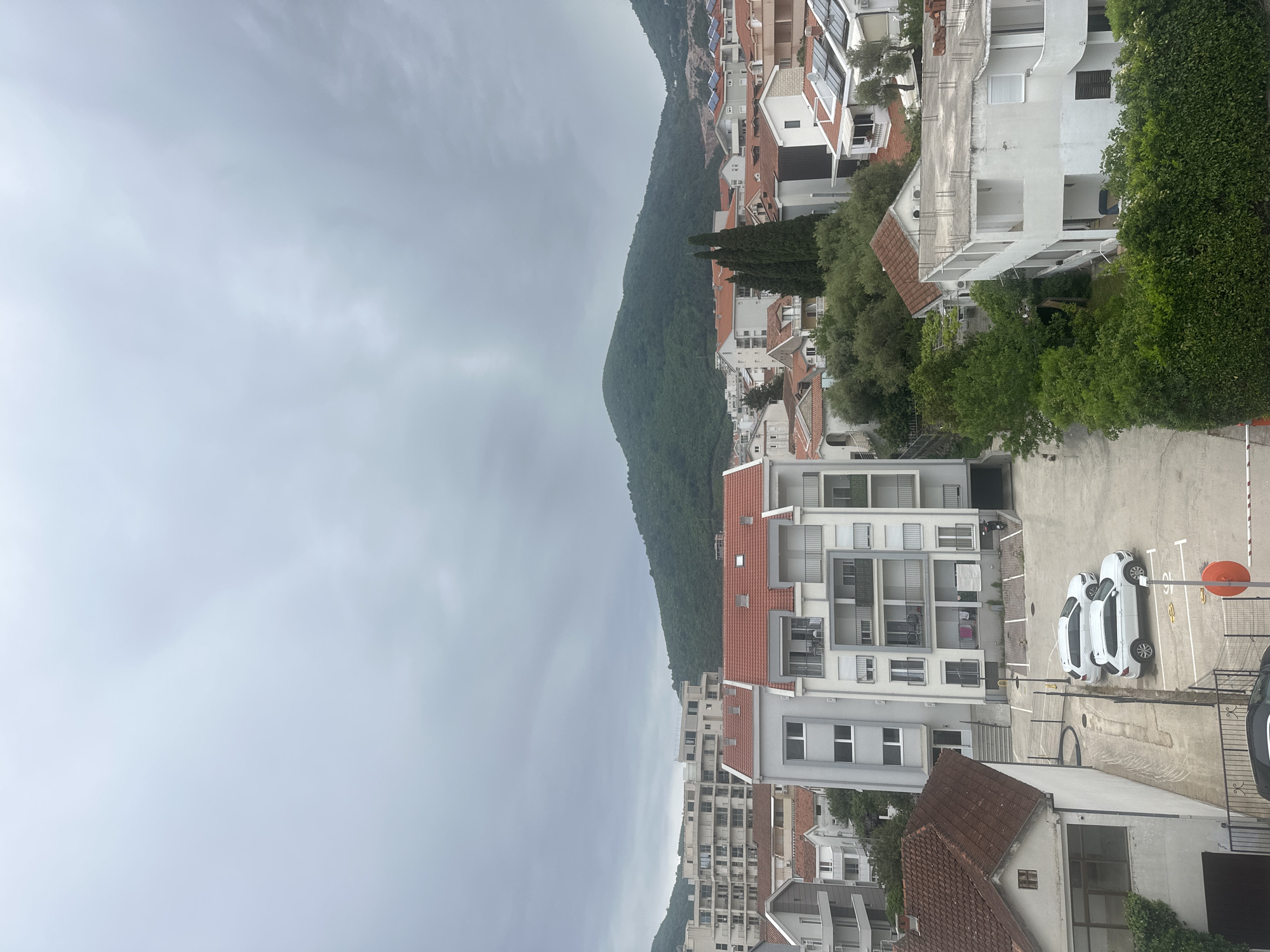 The image depicts an overcast cityscape in Budva, Montenegro. The scene shows an urban area with a mountain backdrop. The architecture consists mainly of white buildings with red-tiled roofs, consistent with the Mediterranean style. In the foreground, there is a parking lot with several white cars. The sky is overcast, creating a muted light across the scene. The buildings appear to be residential, with balconies and windows visible. There are also trees and greenery interspersed among the buildings. A forested mountainside rises in the background. A small, orange sign is visible near the cars in the parking lot.