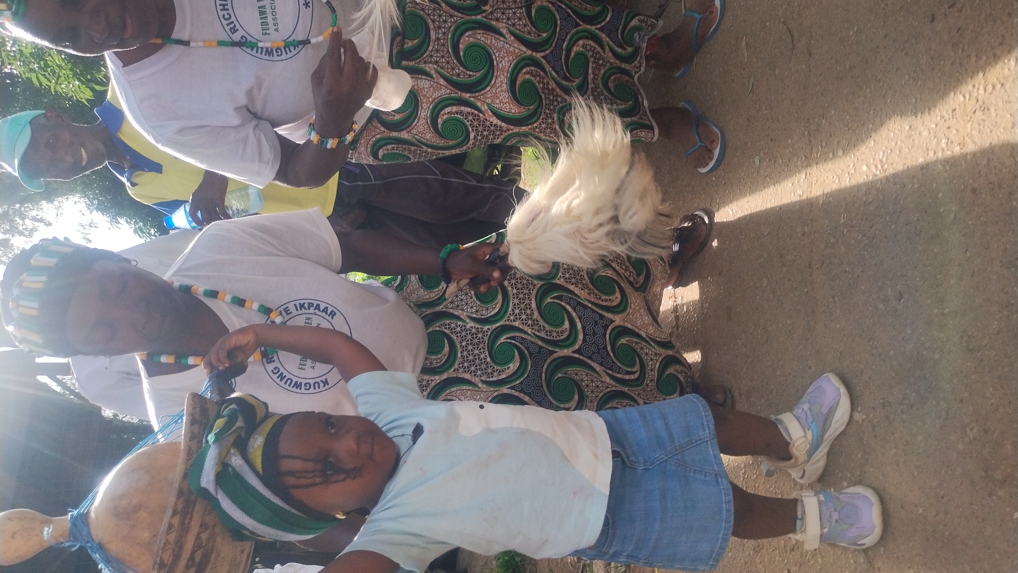 The image depicts an outdoor scene, possibly in Jos, Nigeria, where a group of people are gathered. In the foreground, a young child with braided hair, wearing a light blue t-shirt and a denim skirt, is standing with their arms raised. The child is also wearing light purple and white sneakers. Beside the child, an adult, possibly a woman, is holding a white feathery object, which appears to be a fly whisk or a similar ceremonial item. She is wearing a white t-shirt with a circular logo and a patterned skirt with green and brown swirling designs. Another adult, also in a white t-shirt and patterned skirt, is visible to the right. In the background, a man wearing a yellow shirt and a light blue cap is smiling and holding a plastic bottle of water. The ground appears to be dirt or gravel, and there are shadows cast by sunlight, suggesting it is daytime. The overall atmosphere suggests a cultural event or gathering.