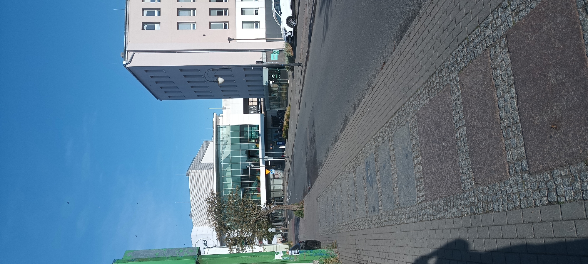 The image depicts a street scene in Świnoujście, Poland, on a clear, sunny day. A modern building with a glass facade, likely a commercial or public establishment, dominates the background. To the right, a multi-story light-colored building with a grid of windows stands adjacent. A white car is parked on the right side of the street. The foreground shows a sidewalk paved with gray bricks and interspersed with rectangular stone slabs framed by cobblestones. A young tree with sparse branches grows from the edge of the sidewalk near the street. The street itself is asphalt and appears wet in patches. The sky is a vibrant blue with a few small white clouds and at least two birds are visible in flight.