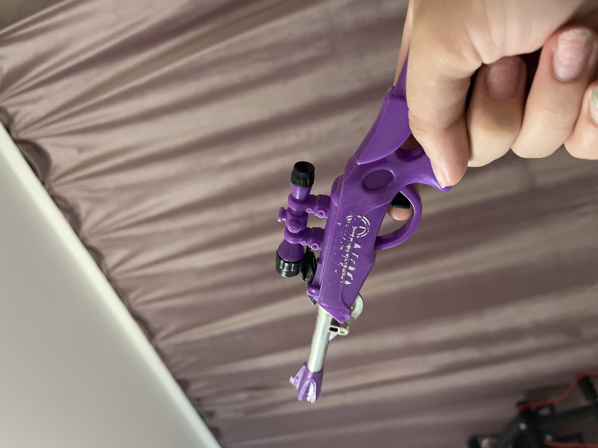A hand holds a purple toy rifle with a scope. The rifle is positioned diagonally across the frame, with the barrel pointing towards the left. The hand is gripping the stock of the rifle, with fingers curled around the trigger area. In the background, a muted pink or mauve curtain is visible, draped in vertical folds. The lighting suggests an indoor setting. There is no indication of the city of Krasnyye Barrikady, Russia in the image.