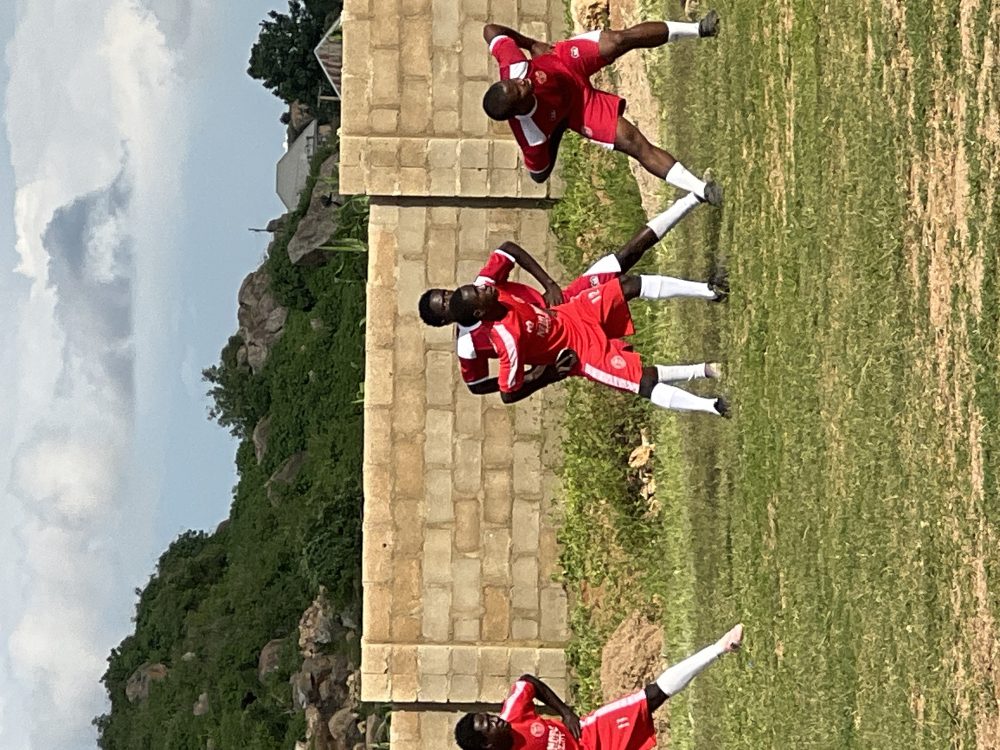 Four young men in red soccer uniforms are performing warm-up exercises on a grassy field in front of a light brown brick wall. The man on the far left, wearing number 11, is stretching his left leg forward and his right leg back. Next to him, a man in the middle is looking to his left, with his arms bent at the elbows. To his right, another man is in a similar stretching pose, with his left leg extended and his right leg bent. The man on the far right is also stretching, with his right leg forward and his left leg back. The background shows a rocky hill covered in green vegetation and a cloudy sky. The scene appears to be outdoors, likely in Jos, Nigeria, given the distinct geological features and vegetation.