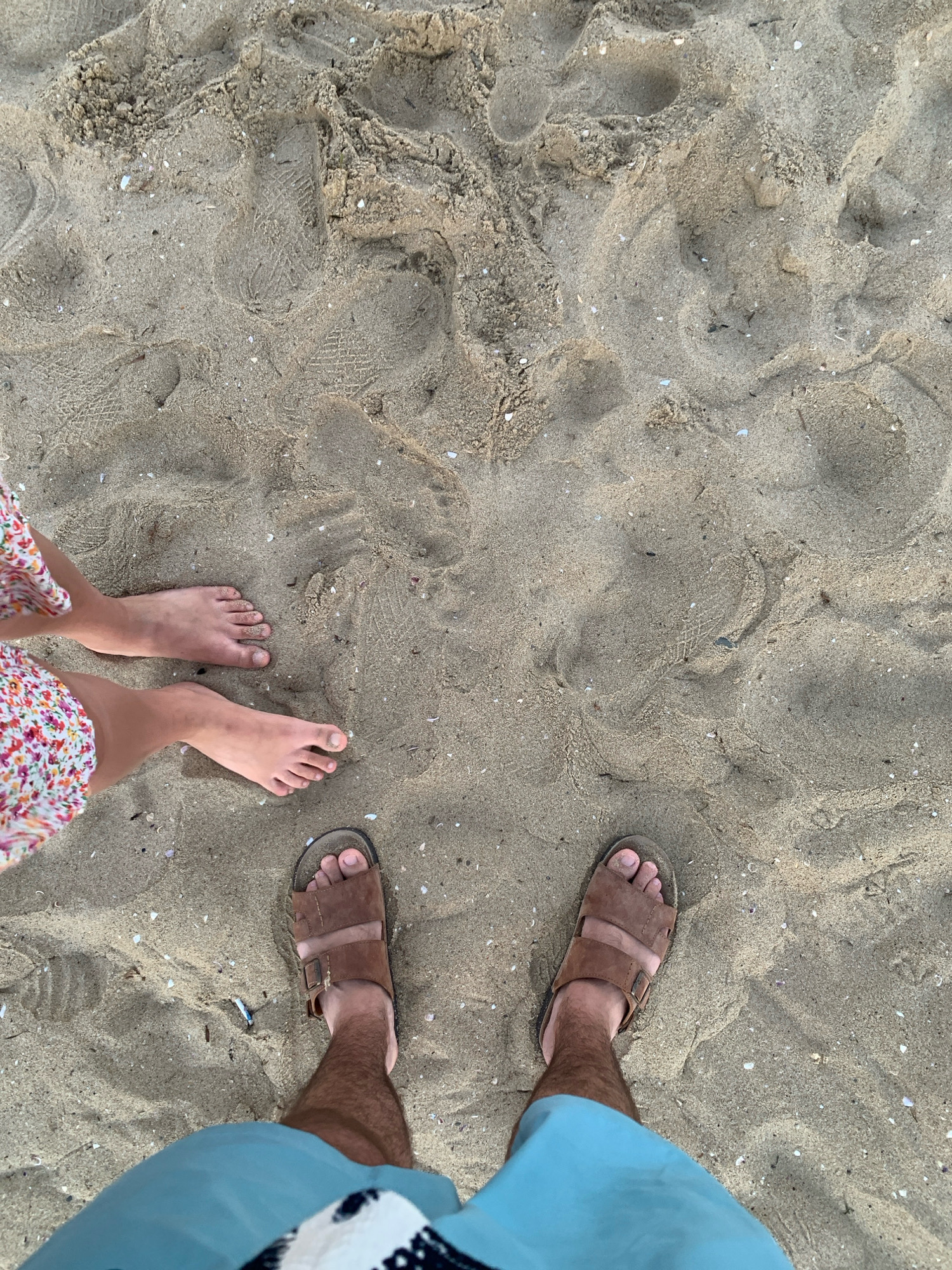 The image shows a top-down view of sandy ground, likely a beach, with two pairs of feet visible. One pair belongs to a person wearing a floral-patterned dress and is barefoot. The other pair belongs to a person wearing light blue shorts and brown sandals. The sand is textured with footprints and small shell fragments. The scene appears to be outdoors, possibly in Nesebar, Bulgaria, given the context. There are no discernible interactions between the individuals depicted.