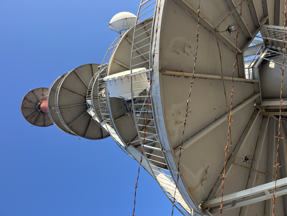 The image displays a tall, grey metallic communication tower situated on Karandila Peak near Sliven, Bulgaria, captured from a low-angle perspective looking upwards against a clear blue sky. The structure comprises multiple spiraling circular platforms, each fitted with white metal railings. Various telecommunication antennas are mounted on these platforms, including white parabolic dish antennas and rectangular panel antennas. Near the apex, a prominent red cylindrical section is visible, capped by a large, flat circular top structure. Several strands of rusted barbed wire are horizontally strung across the foreground and lower tower sections, functioning as a barrier. A caged industrial light fixture hangs suspended beneath one of the lower platforms. Rusting and weathering are evident on the barbed wire and various metallic components of the tower's structure. No individuals are present in the scene.