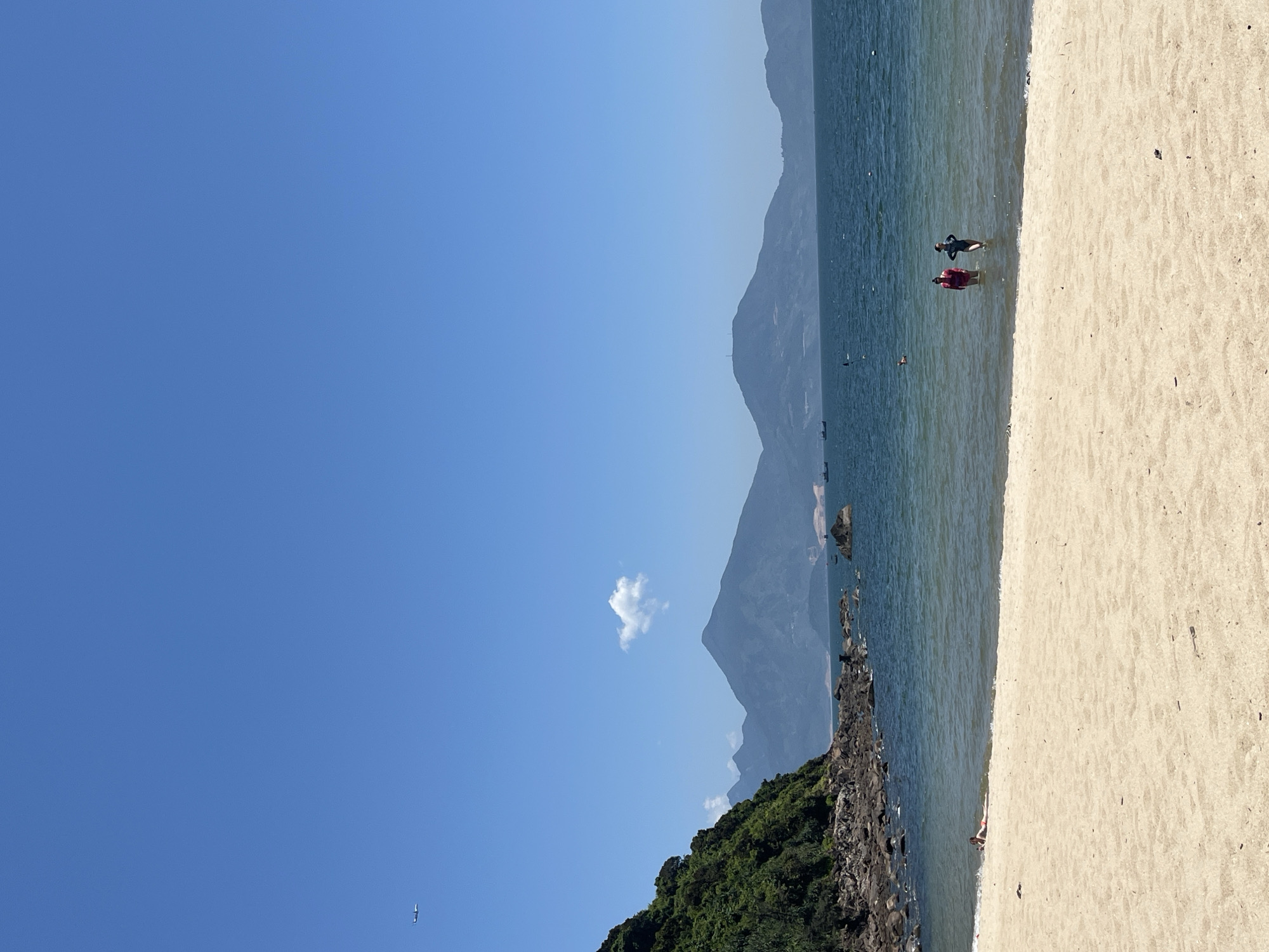 The image depicts a coastal scene with a clear blue sky partially obscured by a small white cloud. In the distance, layered mountains are visible. A rocky shoreline with green vegetation leads to a beach with light-colored sand. Two figures stand in the shallow water at the edge of the beach. An aircraft is visible in the far lower-left corner of the sky. There is no indication that this image is in Đà Nẵng, Vietnam.