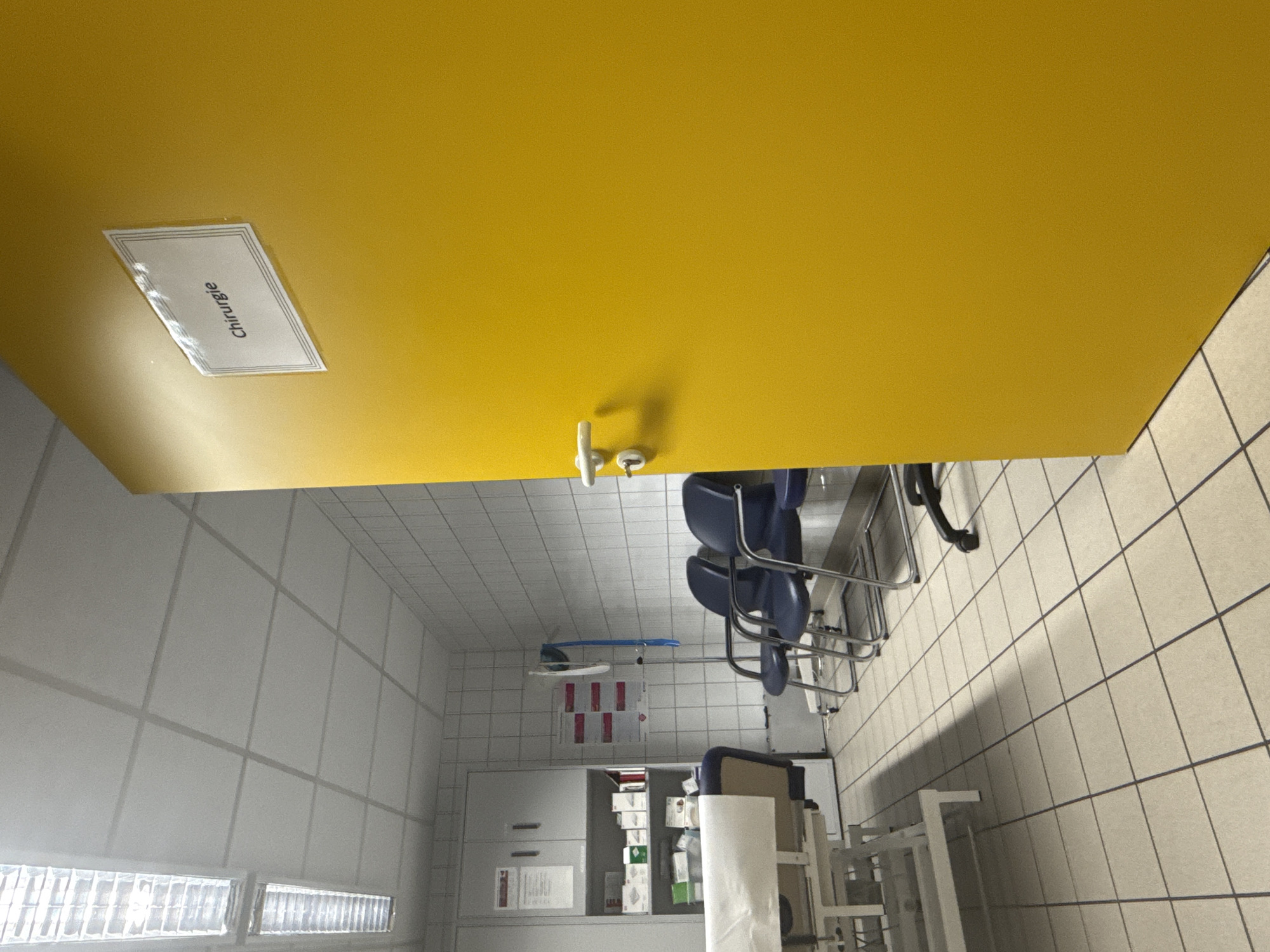 The image depicts a hallway in a medical facility, likely within the town of Walkenried, Germany, given its context. On the right side of the image is a bright yellow door with a silver handle and a small sign labeled "Chirurgie" (Surgery in French or German). To the left of the door, three dark blue chairs with metal frames are arranged, one facing partially towards the door and the other two in a row. In the background, a white tiled wall is visible, with a grey cabinet containing medical supplies to the left. A medical examination table with a white sheet is also present in the left portion of the frame. The floor is tiled with light-colored squares. Overhead, fluorescent lights illuminate the ceiling. The scene appears to be an empty waiting area or corridor outside a surgical department.