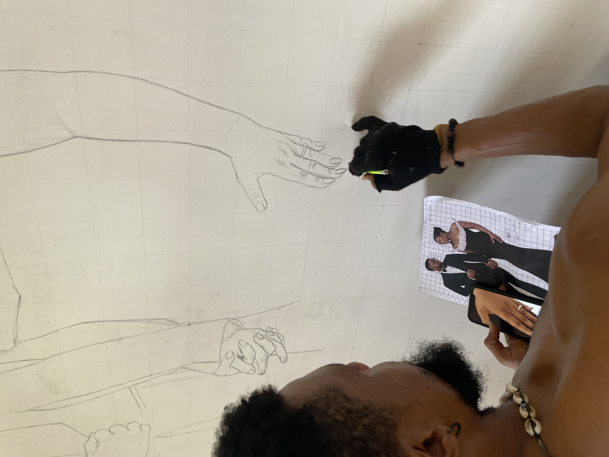 A person is in the process of drawing a large-scale pencil sketch. The drawing depicts intertwined hands and arms on a grid-lined surface. The artist wears a black glove on the hand holding a pencil and refers to a photograph of a formally dressed couple as reference. The photograph is also marked with a grid. The environment appears to be an indoor space with a blank wall. The location is Unknown, Unknown.
