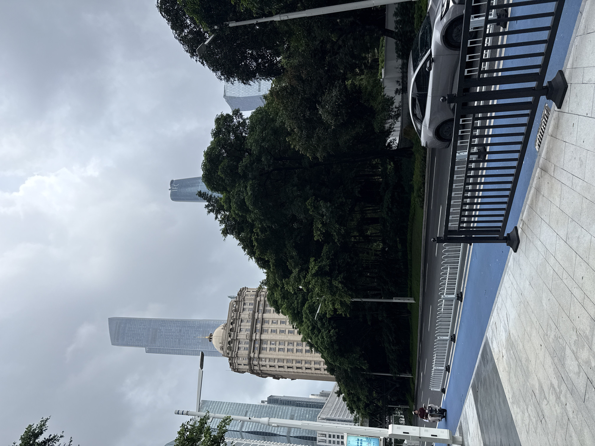 This is a street-level, eye-level, medium shot taken on a cloudy day in Tianhe District, China. The foreground shows a pedestrian walkway made of light gray tiles, with a distinct blue-painted lane for bicycles. A black metal railing separates the blue lane from the street. A silver sedan is visible driving on the street, to the right of the railing. To the left of the blue lane, a person on a scooter is moving away from the camera. The street itself is asphalt and has multiple lanes, marked with white lines. Behind the street, a dense line of dark green trees forms a barrier. Beyond the trees, a large, ornate, beige building with a dome and many windows stands prominently. In the background, several modern skyscrapers of varying heights dominate the skyline. One tall skyscraper to the left of the beige building appears to be made of glass and metal. Another, slightly shorter, futuristic-looking skyscraper is visible to the right, behind the trees. The sky is overcast and a pale gray.