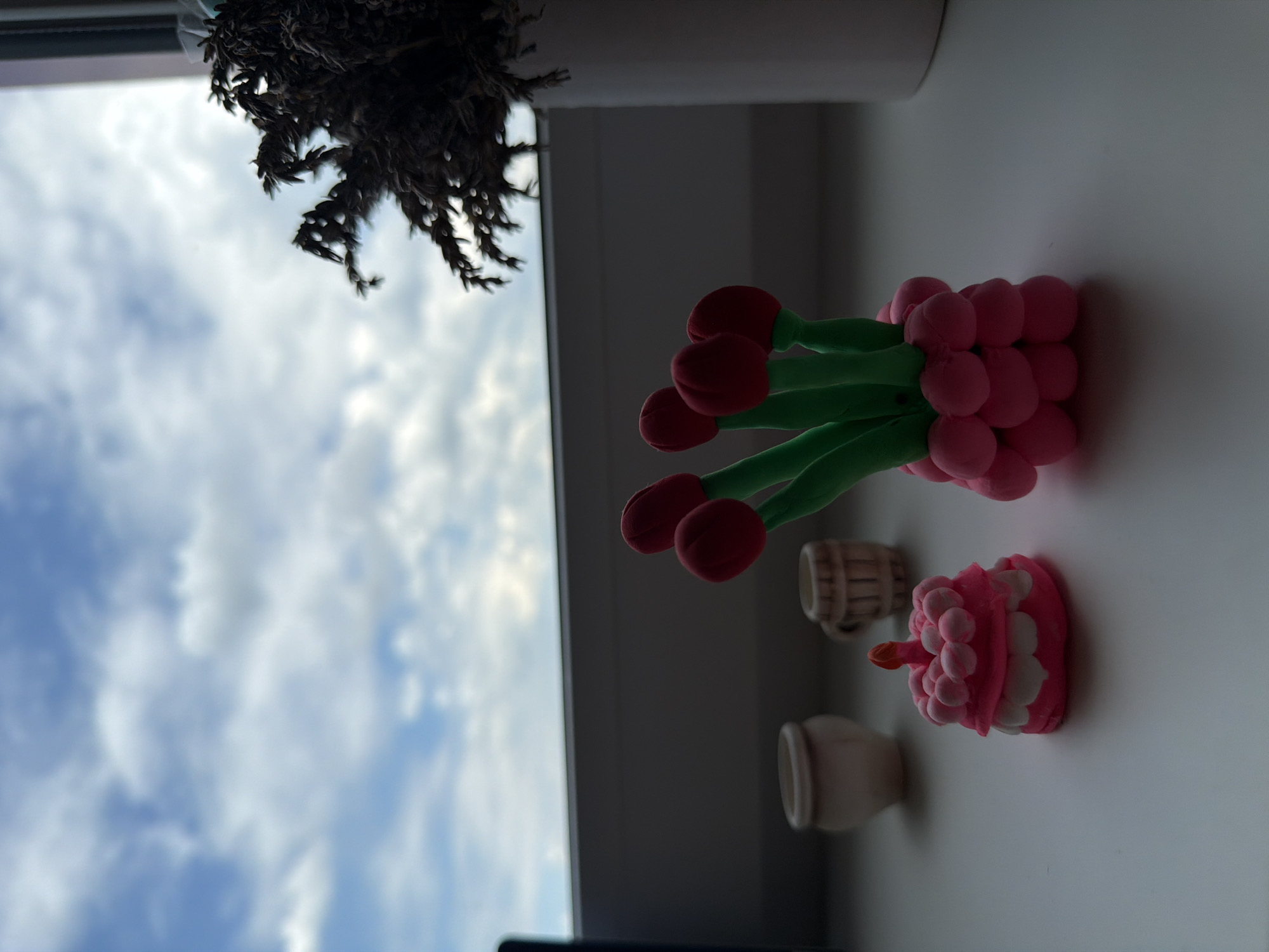 A still life composition is presented on a windowsill. The foreground features a pink and white miniature clay cake with a red candle, and a cluster of red clay tulips with green stems. To the left of the cake are two small, unadorned clay pots. In the background, a white vase holds dried plants. The scene is illuminated by natural light, casting soft shadows. The backdrop is a cloudy sky viewed through a window. The overall setting is indoors, by a window, with the sky visible outside.