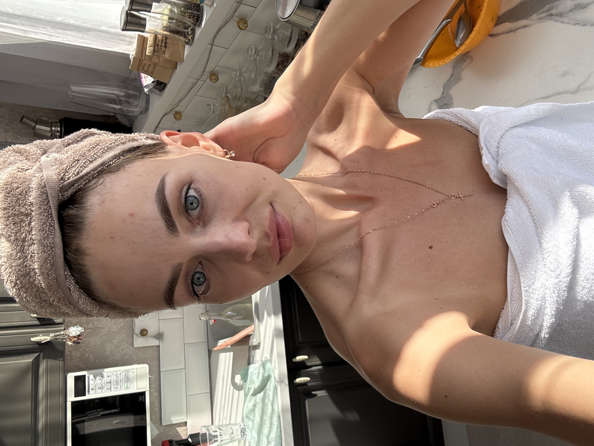 A self-portrait captured indoors depicts an adult female positioned in a residential kitchen in Sochi, Russia. Her head is wrapped in a light brown terry cloth towel, and her body is draped in a white terry cloth towel, covering her shoulders and chest. She has blue eyes, a small red blemish on her forehead, and several small moles are visible on her shoulders and chest. A delicate gold-toned chain necklace with a small cross pendant hangs around her neck. Her right hand (viewer's left) is raised, with fingers lightly touching her ear, which features a small hoop earring with a clear stone.
The kitchen features dark gray upper and lower cabinetry with light-colored circular knobs. A white microwave oven is mounted above a white subway tile backsplash on the left. The countertop is light-colored, possibly patterned marble or quartz. On the counter to the left, a clear glass with liquid, a folded light green cloth, and a red-capped bottle are visible. A sink area with a glass bottle and a light-colored brush or sponge is partially seen.
To the right, a window with sheer white curtains admits strong direct sunlight, casting prominent stripe-like shadows across the countertop and the subject's right arm (viewer's right). The windowsill serves as a display area, holding several clear glass jars containing various contents (e.g., peppercorns), two silver-capped spice grinders, two small paper packets, a clear glass vase, and a metallic thermos. A small hook with decorative charms is visible on the wall above the backsplash. In the bottom right corner, an orange bowl containing a silver spoon and fork rests on the countertop.
