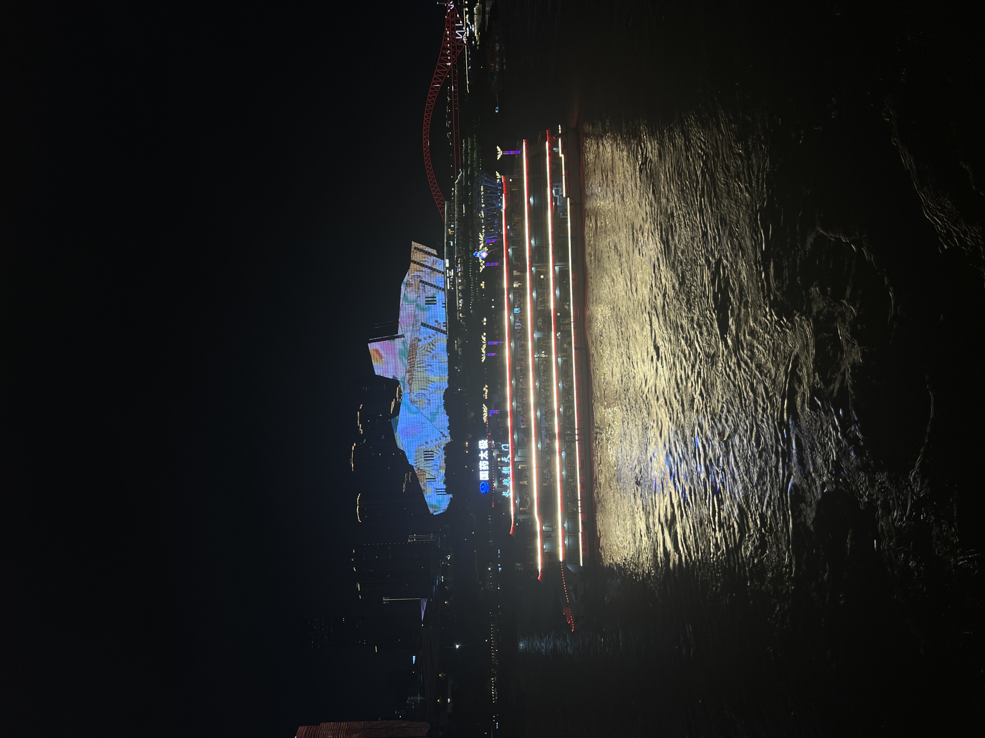 At night, a three-tiered illuminated riverboat is positioned on a dark, rippling body of water. The boat features a red and white lighting scheme along its multiple decks, with numerous small figures visible on board. In the background, a large, modern building is illuminated with a colorful, abstract light show. To the right of this building, a red arched bridge spans the water. The surrounding cityscape is characterized by silhouetted skyscrapers and scattered lights against a dark sky. The scene suggests a vibrant urban nighttime environment.