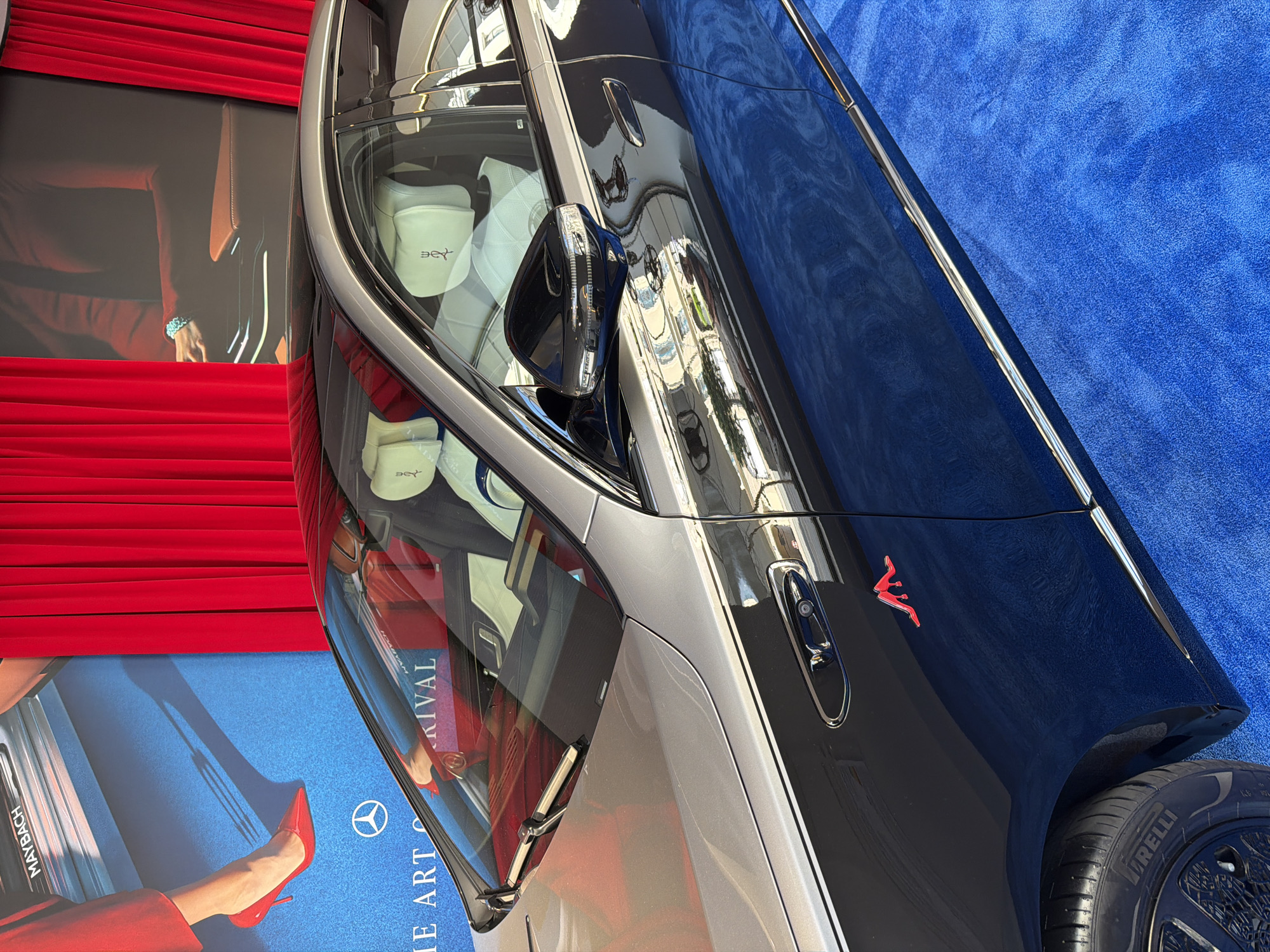 A dark gray and silver Mercedes-Maybach S-Class sedan is parked on a bright blue carpet in front of a red curtain backdrop. The car's interior, with light-colored leather seats, is visible through the windows. A red emblem resembling a stylized figure is affixed to the driver's side door. In the background, a large blue banner displays "MAYBACH" and "THE ART OF" text, along with a red stiletto heel and a Mercedes-Benz logo. The scene appears to be an indoor event or exhibition.