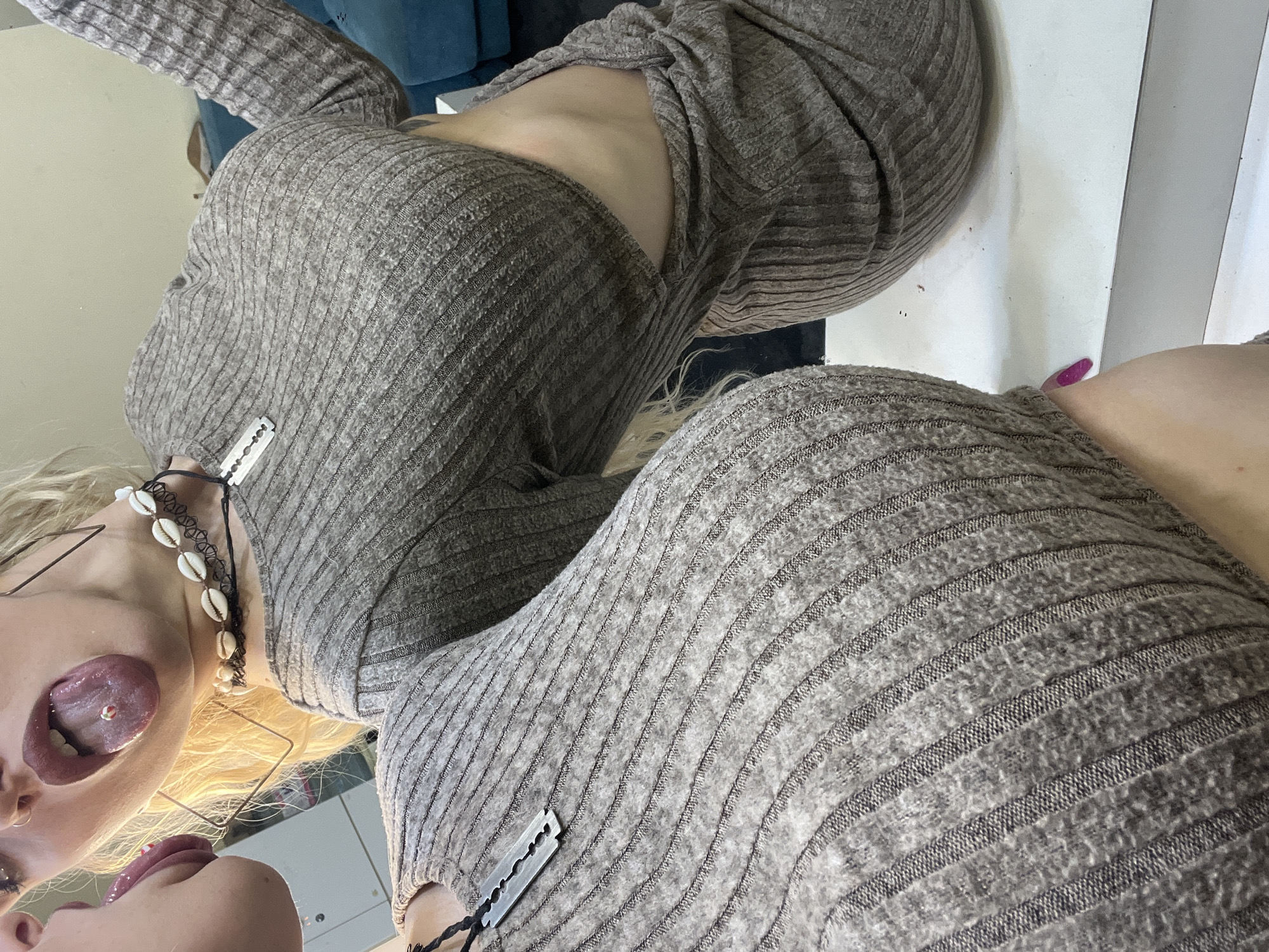 A woman is posing in front of a mirror, wearing a gray ribbed dress and a black seashell necklace. She has blonde hair and a pierced tongue. A razor blade is tucked into the neckline of her dress, both in reality and in the reflection. The location is unknown and there is no indication of it being in Plovdiv, Bulgaria.