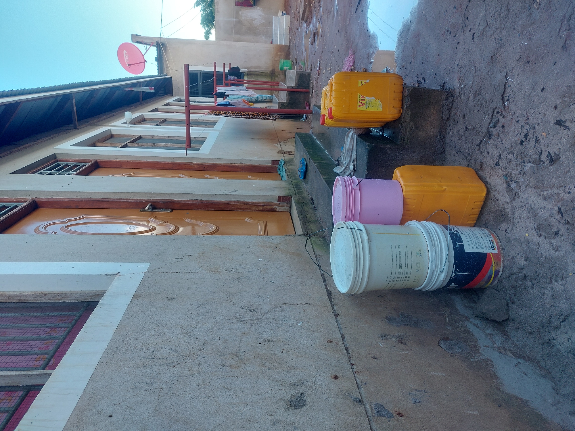 The image depicts a residential area in Dar es-Salaam, Tanzania. The scene is outdoors, likely after rainfall, as evidenced by the wet pavement. The buildings feature a combination of plastered concrete and corrugated metal roofs. Visible are storage containers, including a stack of yellow rectangular jerrycans, a pink bucket, and a paint bucket. A clothesline with clothes hanging to dry is also visible in the background. A red satellite dish is mounted on one of the buildings.