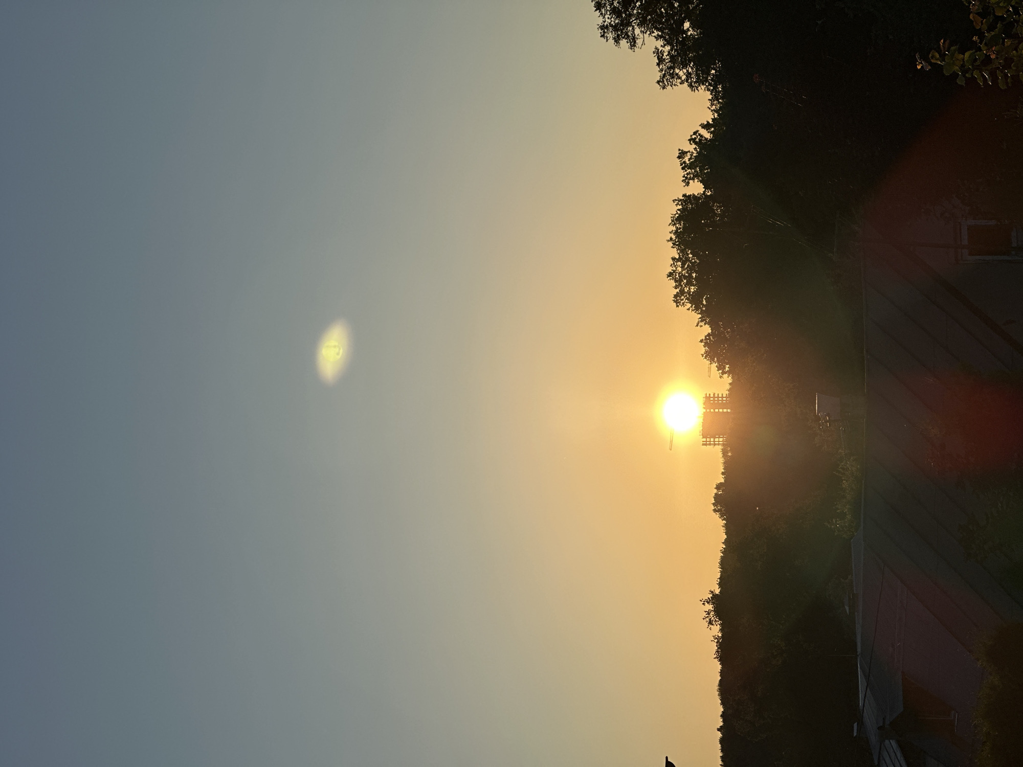 The image depicts a sunset scene in Nizhny Novgorod, Russia. The sun, a bright orb of yellow and orange, is setting near the horizon, casting a warm glow across the sky. The sky transitions from a pale blue on the left to a soft orange on the right, indicating the late hour of the day. Silhouetted against the vibrant sky is a dark line of trees forming a dense forest.
In the foreground, a dark silhouette of what appears to be a bird is visible on a dark surface, possibly a rooftop or a railing. Further to the right, a building with a dark, angled roof is partially visible amongst the trees. The scene is illuminated by the setting sun, creating long shadows and a warm, serene atmosphere. There is no visible text in the image. The overall impression is one of natural beauty and tranquility as day transitions into night.