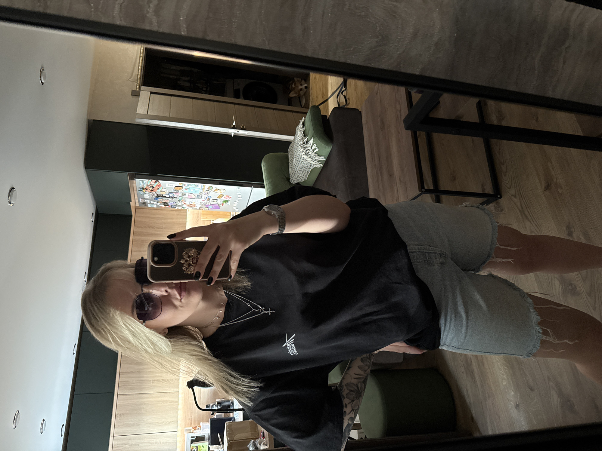 A medium shot depicts a blonde woman in sunglasses taking a mirror selfie. She wears a black T-shirt with white lettering, denim shorts with frayed hems, and a silver wristwatch. Her left arm, visible due to the short sleeve of her shirt, is adorned with a tattoo. She holds a smartphone in her hand, obscuring much of her face.
The reflection shows a part of an apartment interior. A green armchair and a wooden floor are visible. In the background, the kitchen area is equipped with wooden cabinets and a refrigerator covered in magnets. Near the top of the image, a small dog can be seen in a cabinet. The location could not be verified as Krasnoyarsk, Russia using available imagery.