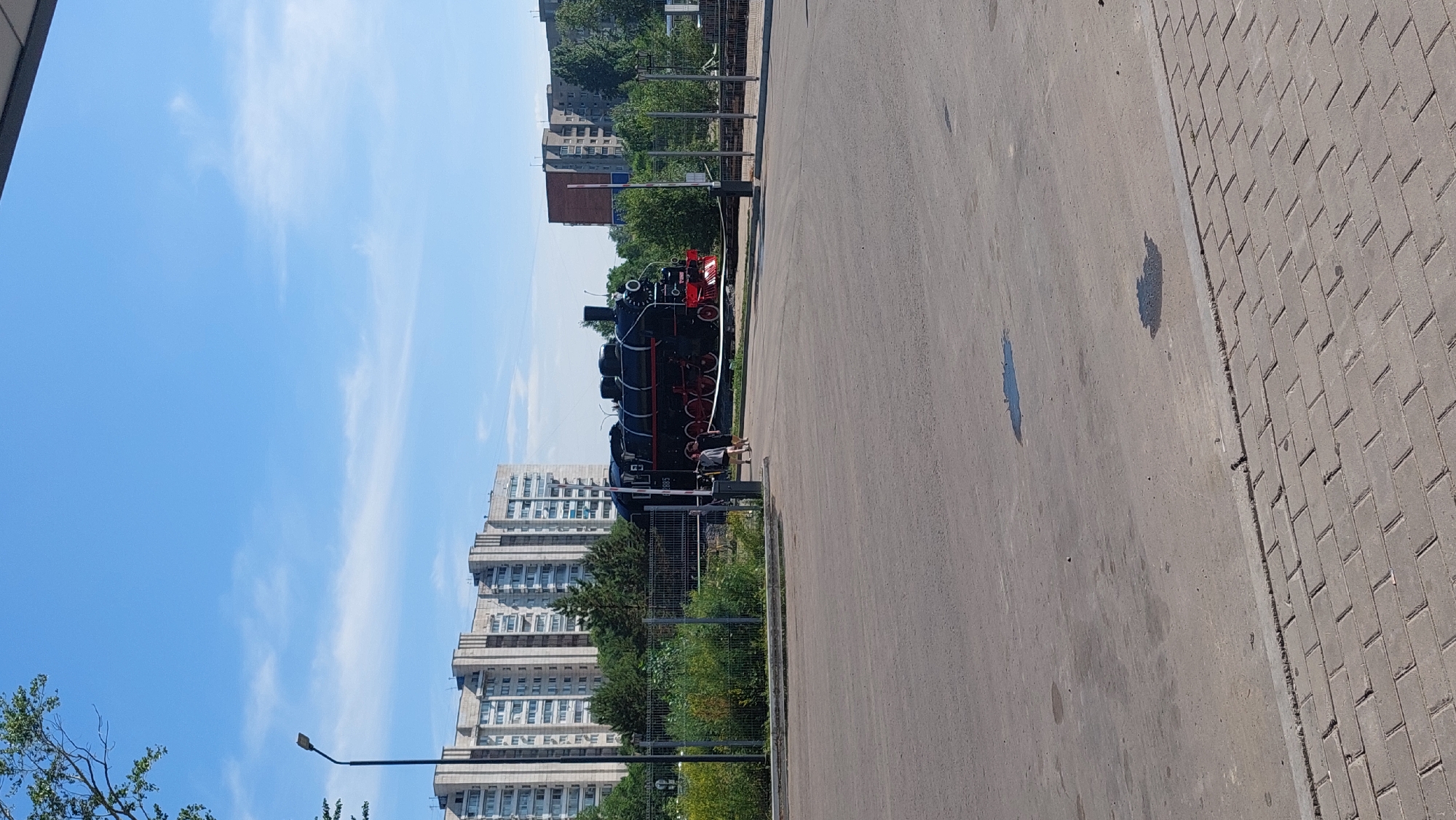 The image depicts a black steam locomotive on display with a building in the background. The locomotive is situated outdoors, with a paved area in the foreground. Two people are visible walking near the locomotive. The sky is blue with scattered clouds. The scene is consistent with a public area or park in the city of Komsomolsk-on-Amur, Russia.