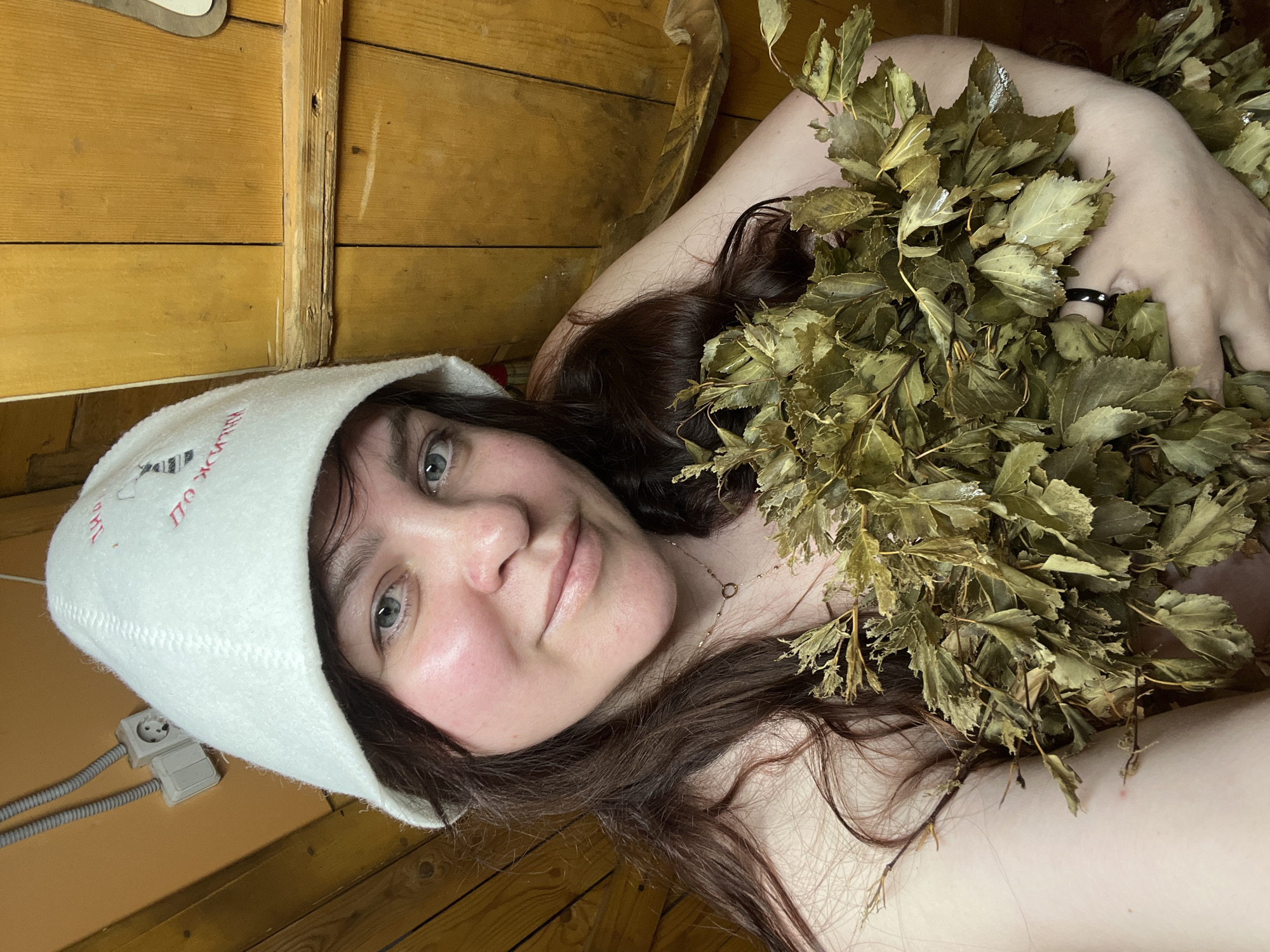The image is a close-up selfie of a woman relaxing in a traditional Russian banya. The setting is an indoor space with warm, light-colored wooden plank walls and what appears to be a wooden ceiling, characteristic of a sauna or bathhouse. Electrical outlets and conduit wiring are visible on the wooden wall in the lower left.
The subject is a woman with long, dark brown hair and light-colored eyes. Her face shows a relaxed, content expression, with a slight smile and visibly flushed cheeks, suggesting she is in a hot environment. She wears a delicate gold necklace with a small circular pendant.
On her head, she wears a white felt sauna hat, which features red embroidered text. Clearly visible on the hat is the Russian phrase "ПО ЖИЗНИ" (PO ZHIZNI), which translates to "FOR LIFE" or "THROUGHOUT LIFE."
She is holding a large, verdant bundle of dried leaves, known as a "venik," commonly used in Russian banyas for whisking the body. The leaves appear to be from birch or oak. A dark-colored ring is partially visible on one of her fingers, partially obscured by the leaves. The overall scene depicts a moment of relaxation and cultural practice within a banya, located in Protasy, Russia.