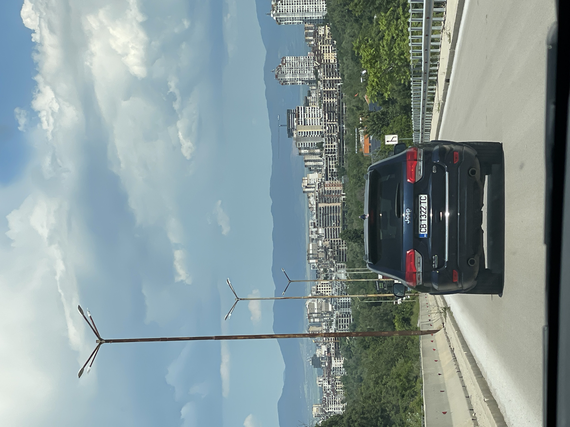 The image depicts a street view in Sofia, Bulgaria, with a dark-colored Jeep Grand Cherokee driving on a multi-lane road. The Jeep has a license plate reading "CB 1322 TC". In the background, a cityscape is visible with numerous apartment buildings and taller high-rises, indicating an urban setting. Beyond the city, a range of mountains forms the horizon. Several streetlights are present along the road, one notably tall and with multiple lamps. The sky is partly cloudy, with patches of blue sky visible. Green trees and foliage line the sides of the road and the surrounding landscape, suggesting the presence of parks or natural areas within or near the city. The road appears to be an elevated section or a bridge, with concrete barriers and metal railings on either side. The perspective is from a vehicle traveling behind the Jeep.