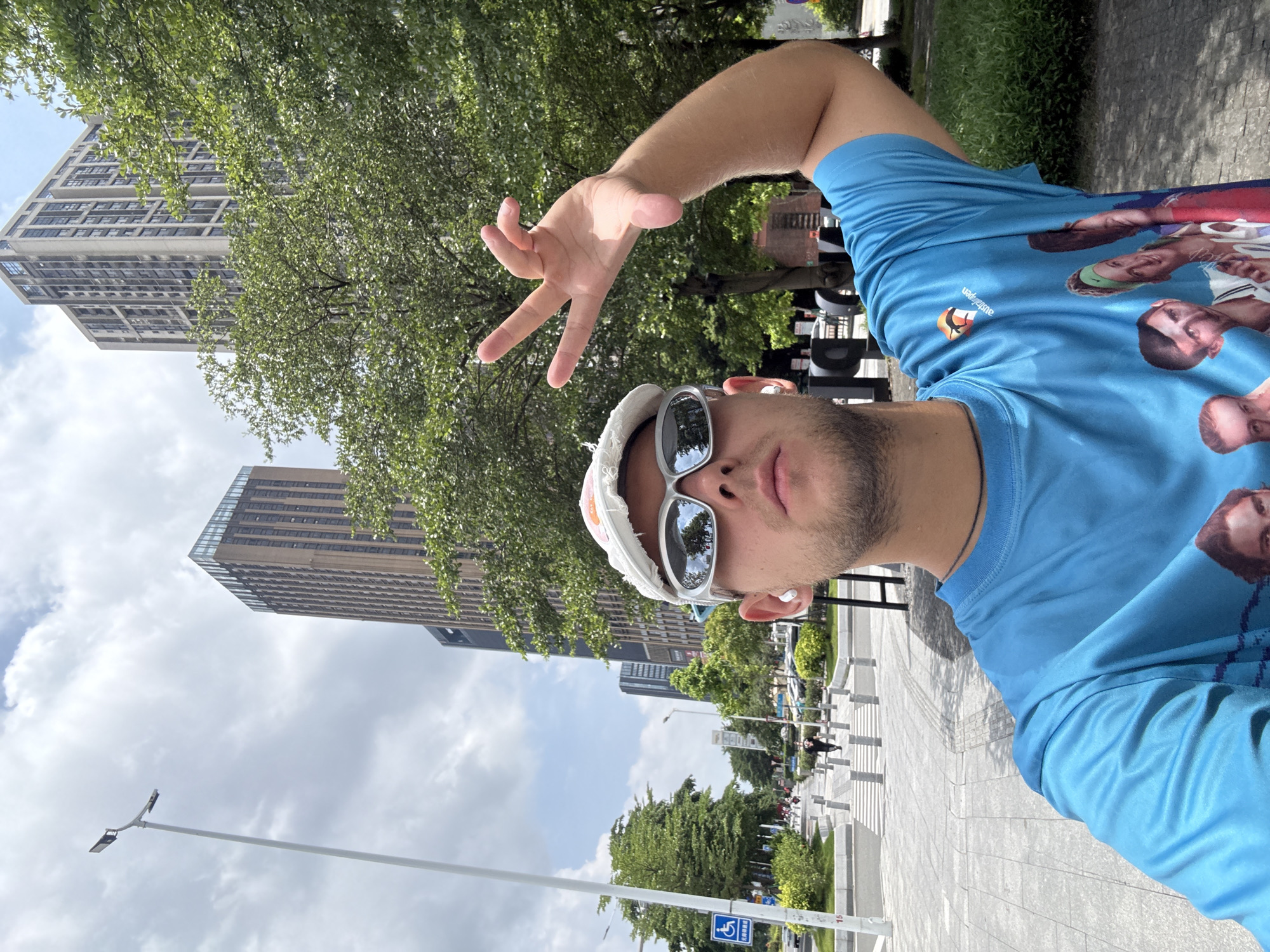 The image is a selfie taken outdoors in Haizhu District, China. The foreground features a person wearing a white cap with orange accents, reflective sunglasses, and a bright blue t-shirt with a graphic print of several faces. The person is also wearing white earbuds and has their right arm raised, with their hand making a "V" gesture. In the background, there are modern tall buildings, lush green trees, and a paved walkway with bollards and streetlights. A blue sign with white text and a wheelchair symbol is visible on the left side of the walkway. The sky is partly cloudy.