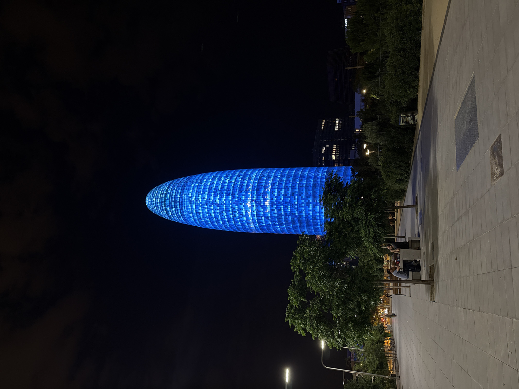 The image captures a night-time view of the Agbar Tower in Barcelona, Spain. The distinctive skyscraper, illuminated in a vibrant blue, dominates the skyline. In the foreground, a tree-lined pedestrian walkway is visible, paved with light-colored square tiles. Several people are seated at picnic tables under the trees, engaged in conversation. Streetlights cast a warm glow on the walkway, contrasting with the cool blue of the tower. Other buildings and greenery are present in the background, indicating an urban park or public space. The sky is dark, with a few scattered clouds, and the overall atmosphere is calm and urban.