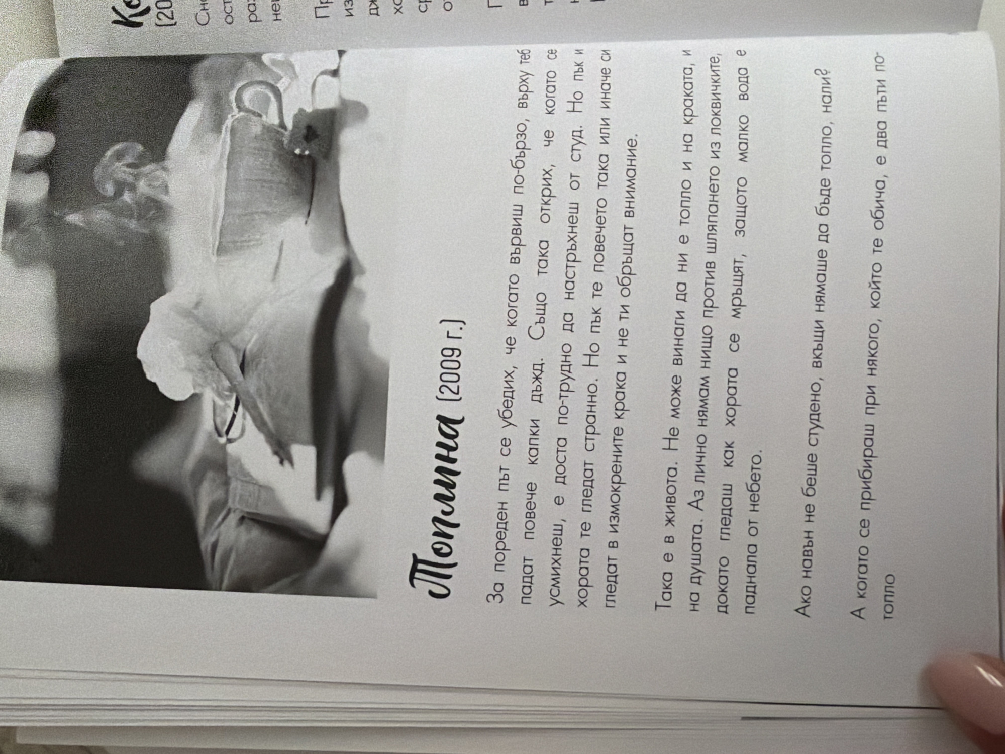 The image shows a close-up of an open book with text and an illustration. The illustration is a black and white photograph of a steaming cup of coffee or tea placed on what appears to be a book or folded cloth. The steam rises from the cup in an artistic manner. The text is in Bulgarian. The left page of the book has a title in stylized font: "Топлина (2009 г.)". Below the title are several paragraphs of text. The right page of the book contains some text, including the start of a word "Ко" and a year in parentheses "(2009)". The image is a photograph taken from above, focusing on the book. There are no identifiable landmarks or features that would place the scene in Pazardzhik, Bulgaria. The image provides no information about the city of Pazardzhik.