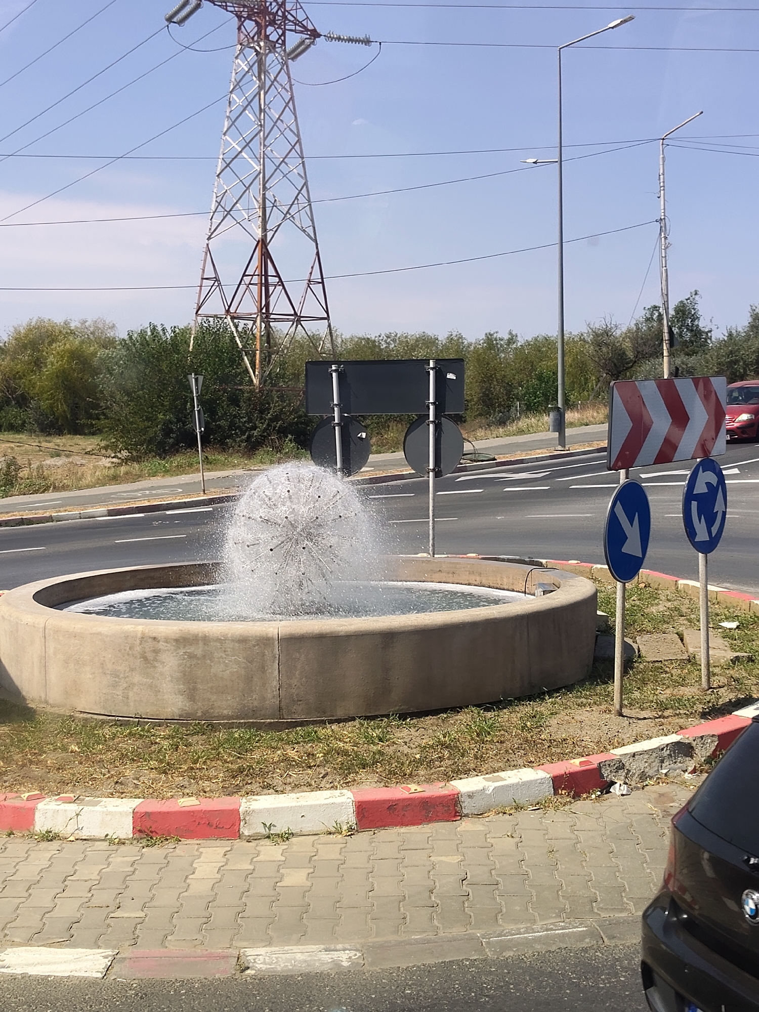 A wide shot captures an outdoor scene on a bright day, featuring a roundabout with a spherical water fountain at its center. The fountain is a focal point, with water spraying upwards and outwards, creating a mist. The roundabout is bordered by a concrete curb painted with alternating red and white stripes, and beyond that, a paved sidewalk.
In the background, a large red and white chevron sign is mounted on a pole, indicating a turn. Below it, two blue circular traffic signs are visible: one with a white arrow pointing left, and another depicting a circular arrow, signifying a roundabout. A dark grey car is partially visible on the right side of the frame, parked near the sidewalk.
The environment is characterized by a clear blue sky with a few wispy clouds. A tall, lattice-like electricity pylon stands prominently in the left background, with power lines stretching across the sky. Streetlights are also present, with one tall, grey pole situated to the right of the pylon and another shorter one further back. Trees and foliage line the sides of the road, suggesting a somewhat rural or suburban setting. The road itself has white lane markings. The overall impression is one of a functional traffic intersection with a decorative element.