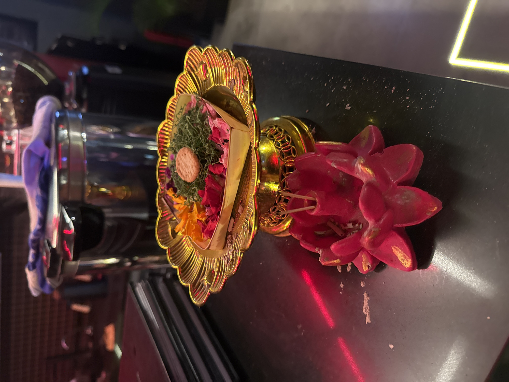The image captures a Balinese offering, likely found in the Kuta Selatan area of Indonesia. The offering consists of a golden, intricately designed tray holding a small collection of organic materials including moss, petals (red and yellow), and what appears to be a biscuit or wafer. Adjacent to the tray is a small arrangement of artificial red flowers. The objects rest on a dark surface that shows signs of wear and residue. A blurred backdrop features a reflective surface, indicating a contemporary interior setting.