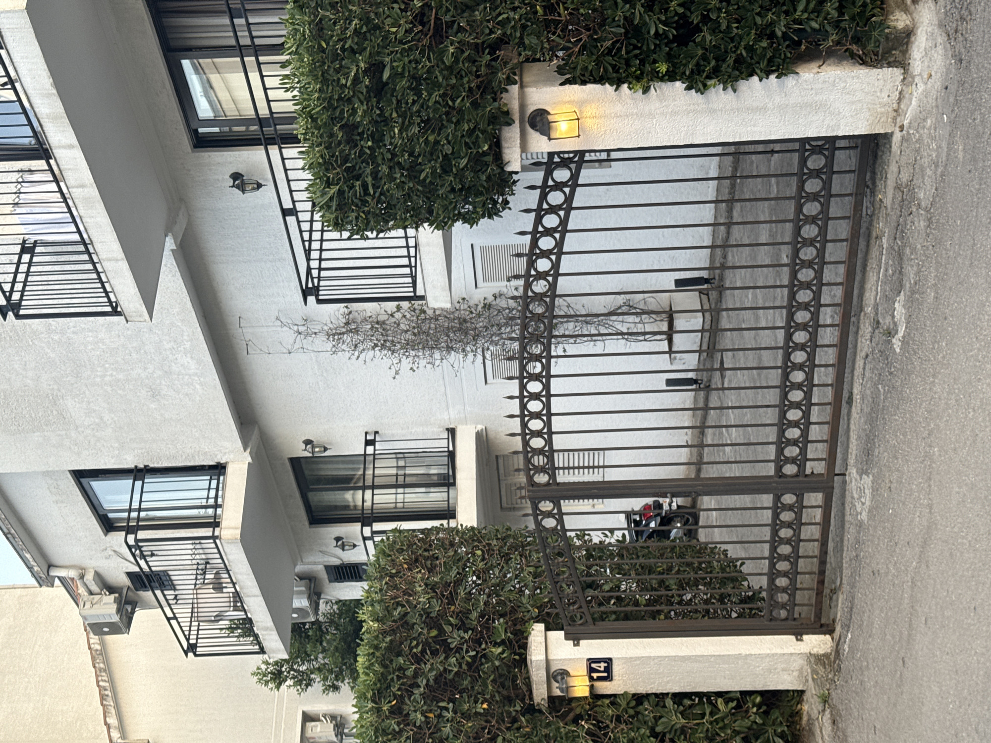 A daytime shot taken in Budva, Montenegro, shows the exterior of a building featuring a white facade, dark-framed windows, and iron balconies. A wrought-iron gate, adorned with circular designs, stands at the entrance to the property. A small tree grows in front of the building. A trimmed bush partially obscures the gate on either side. A small sign displaying the number "14" is visible on a pillar near the gate. A parked motorcycle is visible through the gate. The building is located on an asphalt road.