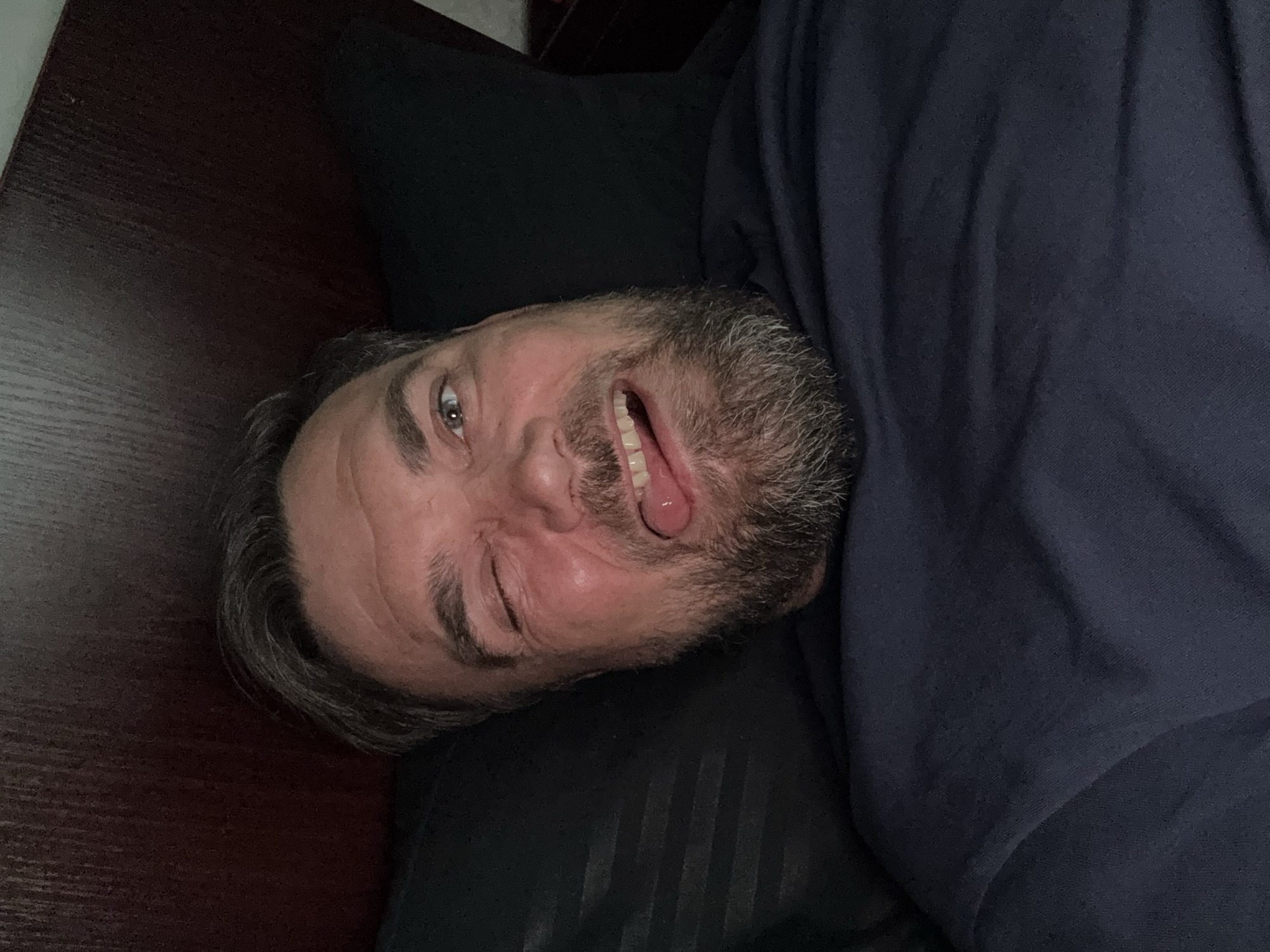 A man is lying on a bed, his head propped up on a pillow. He is looking at the camera and sticking his tongue out. He is wearing a navy blue t-shirt. The background is a dark headboard of a bed.