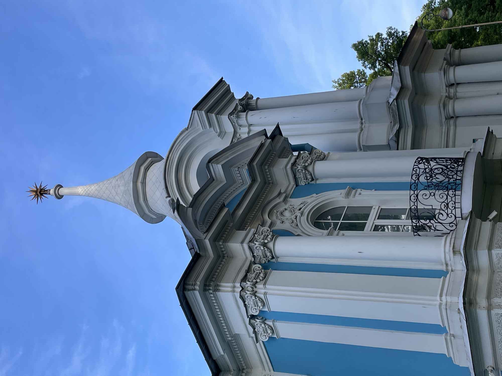 This image displays a low-angle view of an architectural structure located within the Peter and Paul Fortress in Saint Petersburg, Russia. The building is predominantly white and bright blue, featuring classical architectural elements under a clear blue sky.
The main body of the building exhibits vertical stripes formed by bright blue wall sections alternating with white pilasters. These white pilasters are topped with ornate Corinthian capitals, which support an elaborate, multi-layered white cornice adorned with decorative moldings, including dentils. Rising from an upper section of the structure is a slender, conical white spire, textured with a subtle pattern, culminating in a prominent, gilded, multi-pointed star finial at its peak. A small bird is perched on a white ledge at the base of the spire on the right side.
On the visible right section of the building, an arched window with white frames and multiple panes is present, positioned above a small, ornamental black wrought-iron balcony featuring intricate scrolling patterns. To the right of this, a white column with a Corinthian capital extends upwards. In the background on the right, a cluster of dark green trees is visible. A vintage-style streetlamp with a spherical, translucent light fixture is partially visible on the far right, extending from behind the building. The sky is a vibrant, uniform blue with minimal, faint wisps of white cloud.