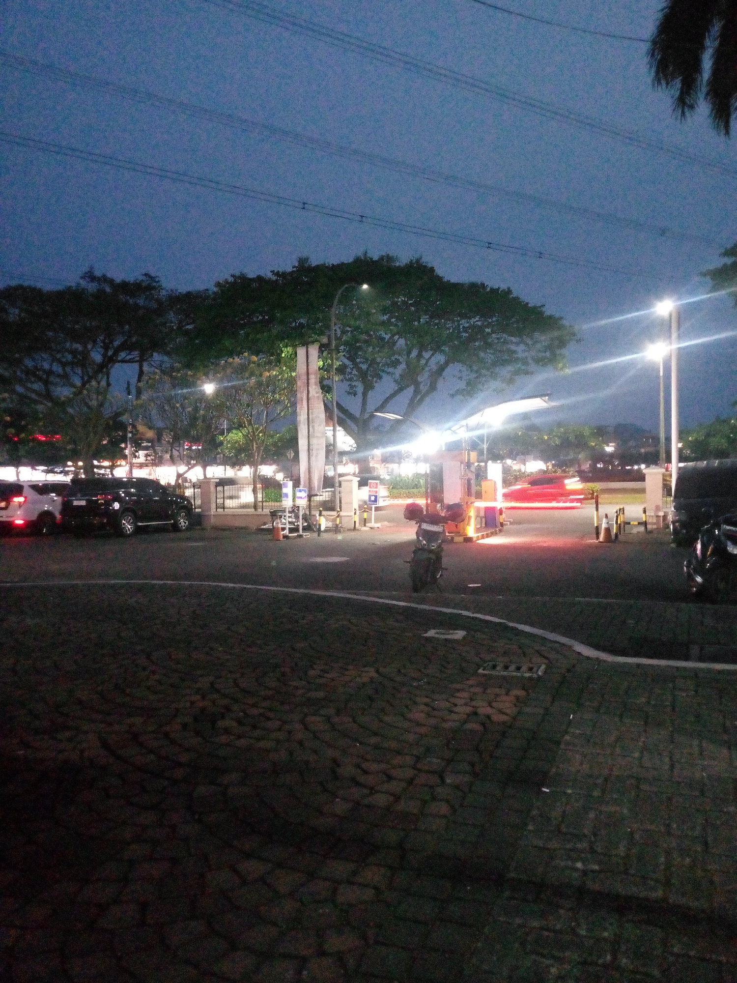 The image depicts a street scene at dusk or night in Cikupa, Indonesia. The foreground is a cobblestone-like paved area. In the middle ground, several vehicles are visible, including a white car and a dark SUV parked near what appears to be a toll booth or entrance to a facility. A motorcycle is also parked in front of the structure. Bright streetlights illuminate the area, casting reflections and creating light streaks from moving vehicles, most notably a red blur suggesting a car in motion. Tall trees with dense foliage line the background, and power lines are visible against the twilight sky. Buildings and more trees are faintly discernible in the distance. The overall impression is of a moderately busy urban or semi-urban environment at the end of the day.