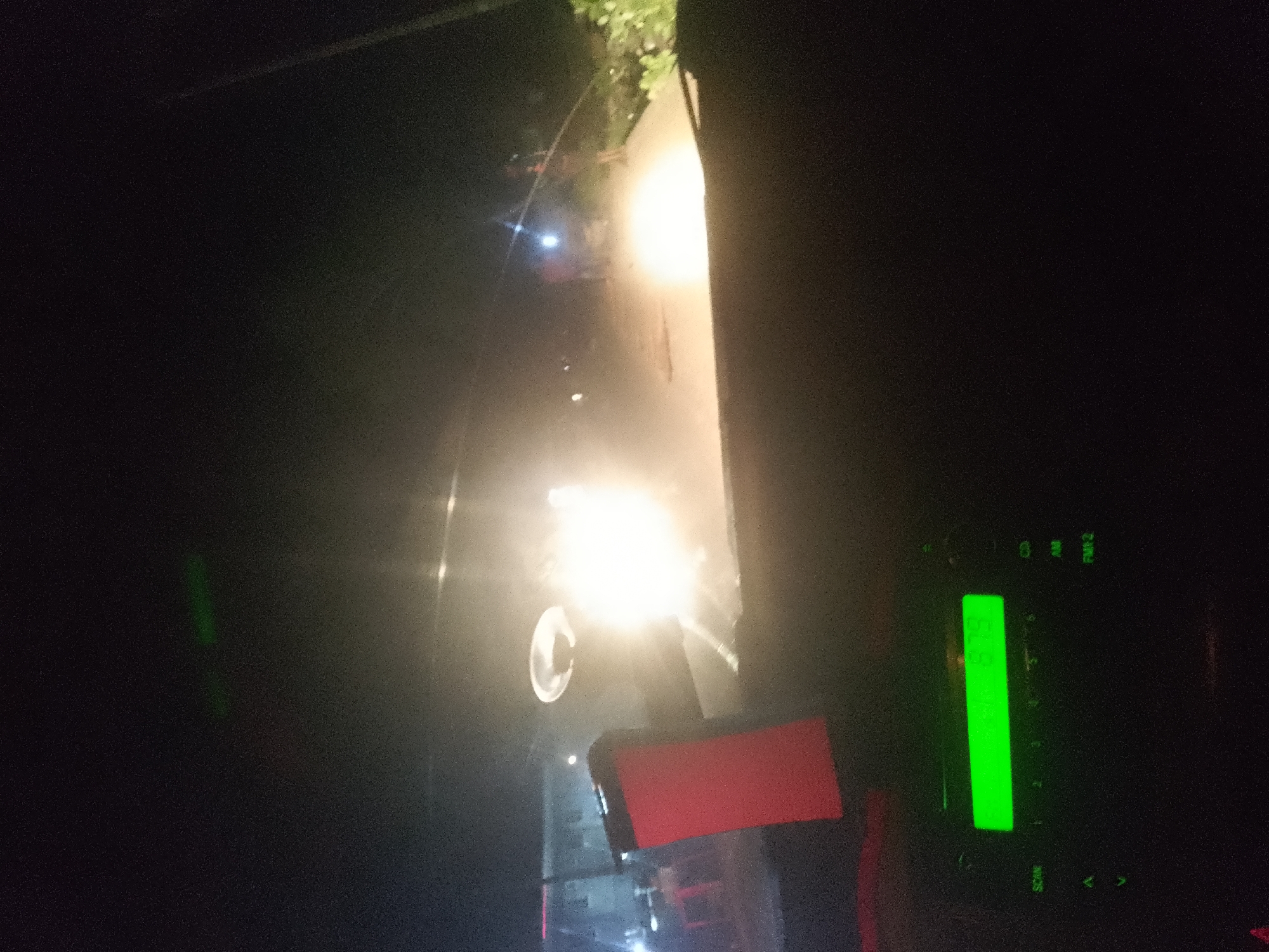 The image shows a dark, outdoor scene at night, likely from inside a vehicle. In the foreground, the right side of the frame displays a car's dashboard with a bright green digital display showing "103.95" and some buttons labeled "AM", "FM 2", "SCAN", and directional arrows. A bright red object, possibly a phone mount or a piece of trim, is also visible on the dashboard. The background is illuminated by intense headlights from an unseen source, creating a hazy glow that washes out details. Some foliage or trees are dimly visible at the top of the frame, and a pale wall or structure is illuminated on the right. The overall impression is of a vehicle moving through a dimly lit urban or suburban environment at night.