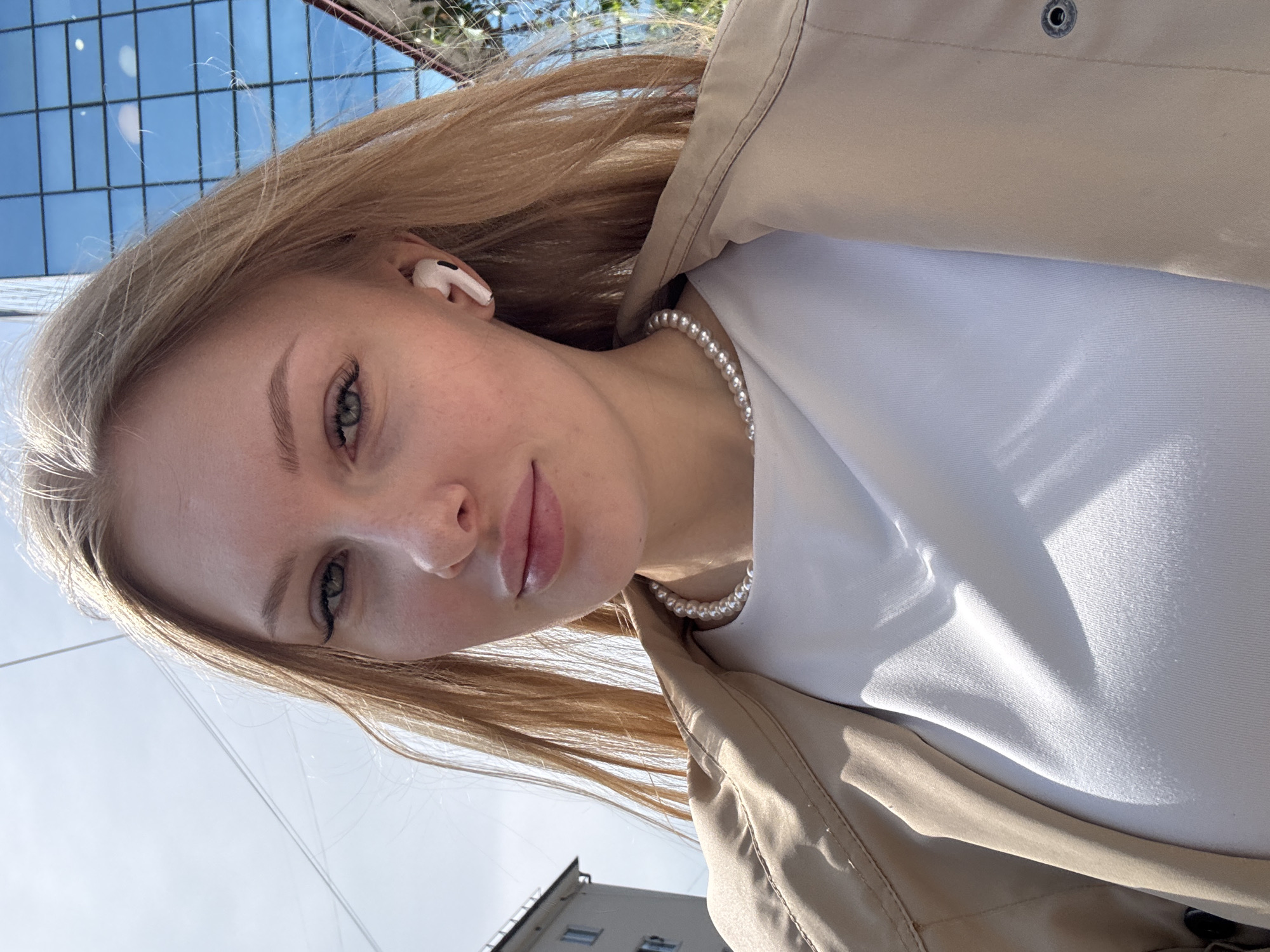 A young woman with light blonde or light brown hair and green eyes is featured in a close-up, upward-angled selfie. She is looking directly at the camera with a gentle, slightly smiling expression. Her lips appear to have a natural or light pink gloss.
She is wearing a white earbud (resembling an Apple AirPod) in her right ear. Around her neck, she has a white pearl necklace. She is dressed in a white top or t-shirt, partially covered by a light-colored, possibly beige or tan, jacket or trench coat with a visible button or snap on the collar/lapel area.
The background suggests an outdoor, urban setting during bright daylight. Above and to her left, a clear blue sky is visible, with what appears to be reflections of square window panes from a building or overhead structure. Below and to her right, a portion of a building with windows can be seen, hinting at architecture consistent with a city like Perm, Russia. The sunlight casts subtle shadows, indicating a sunny day. No visible text is present in the image.