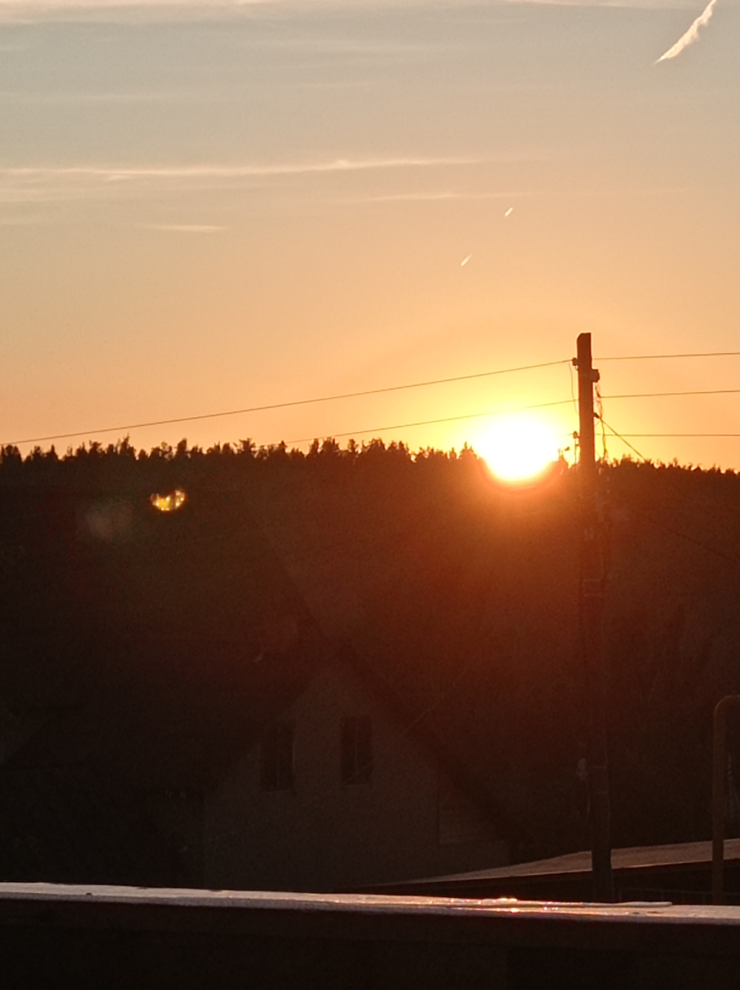 The image shows a sunset over a dark, silhouetted forest with a power line pole in the foreground. The sky is a gradient of orange and yellow, with a few wispy clouds. The sun is a bright orb just above the treeline, casting a warm glow. There appears to be a building or structure partially visible through the trees, with windows that reflect the sunlight. The overall scene is peaceful and serene, capturing the beauty of a natural landscape. The location is in Degtyarsk, Russia, a city known for its industrial history and surrounding natural beauty.