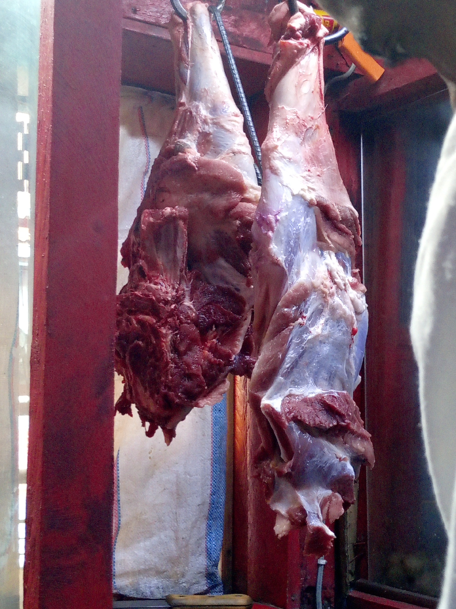 Two pieces of raw meat hang from metal hooks in a market setting. The meat appears to be ungulate carcasses, partially skinned and butchered. Red painted wooden beams frame the scene, suggesting a market stall. In the background is a white cloth or bag. The setting suggests a butcher shop, possibly in Nairobi, Kenya, where such open-air markets are common. The image contains no visible people, interactions, or actions.