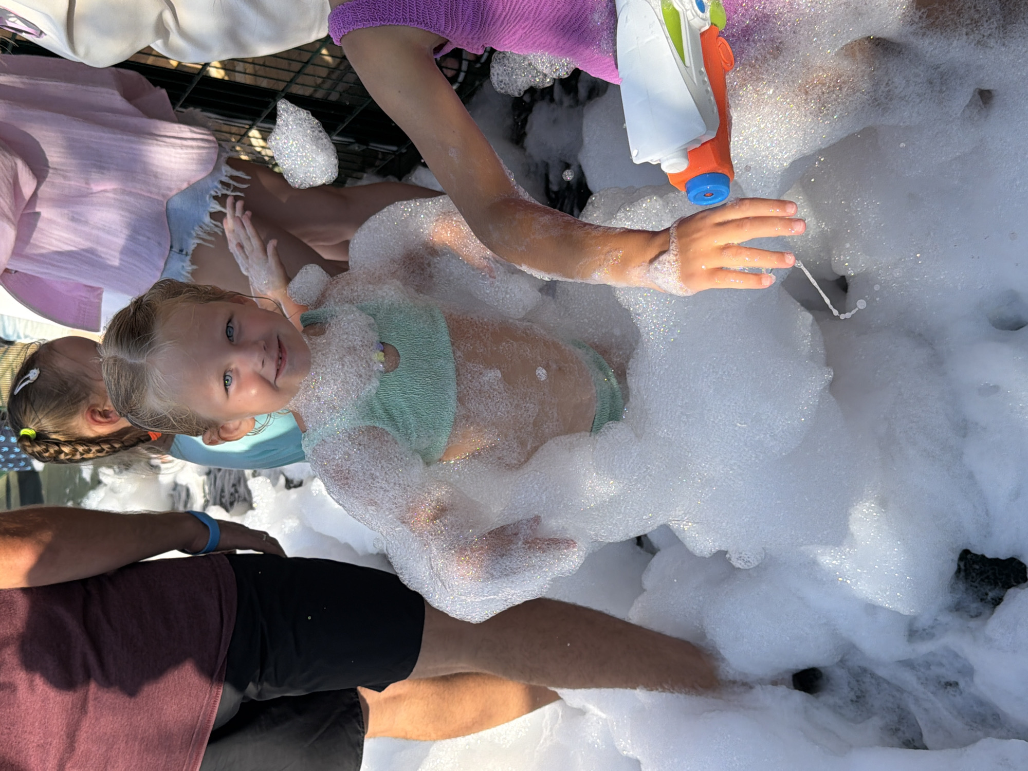 In this overhead shot, a young girl with blue eyes and blonde hair is the central subject, immersed in a sea of white foam. She wears a mint green bikini top and appears to be enjoying herself, her hands spread wide in the bubbles. To her left, another child, possibly a sibling, is partially visible, with braided hair adorned by a yellow tie. In the background, a person in a purple top and a pink shirt can be seen, along with a white and orange water gun. The scene suggests a playful outdoor activity, possibly a foam party or a water-based celebration, given the presence of the water gun and the copious amount of foam. The lighting indicates it's daytime, likely a sunny day, judging by the bright reflections on the foam. The overall mood is cheerful and carefree, with the children clearly engaged in a fun-filled moment.