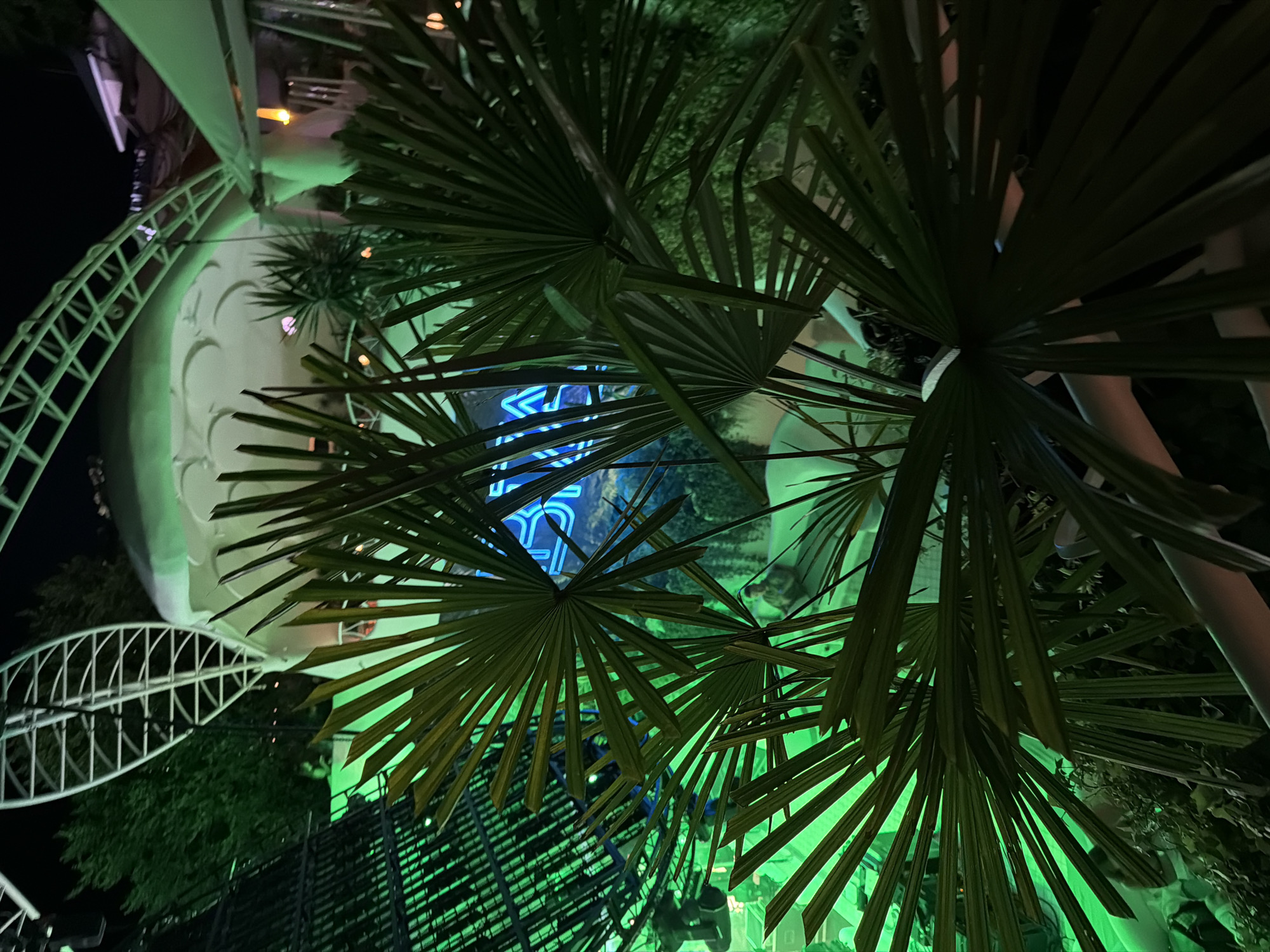The image depicts a neon sign that spells out "ODESA" in blue, set against a backdrop of lush green foliage. The sign is positioned within an outdoor establishment, likely a restaurant or club, illuminated by vibrant green lighting that casts a dramatic glow on the surrounding palm fronds. The architecture suggests a contemporary design, with a white domed structure and a metal canopy visible in the background. The overall ambiance is one of nightlife and urban entertainment. The presence of the neon sign clearly identifies the location as Odesa, Ukraine.