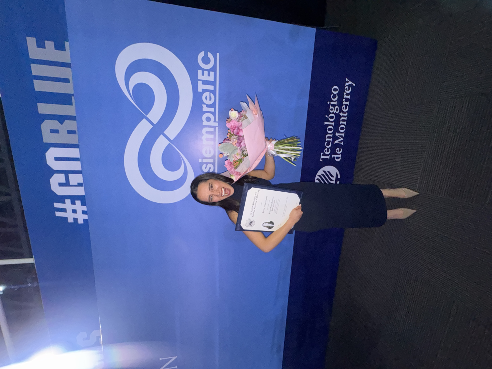 A young woman is posing for a photo in front of a Tecnológico de Monterrey banner, likely at the Puebla, Mexico campus. She is smiling, holding a bouquet of flowers and a diploma. The banner is blue with white lettering that reads "#GOBLUE" and "siempreTEC". The Tecnológico de Monterrey logo is also visible on the right side of the banner. The woman is wearing a dark dress and light-colored heels. The background is a dark, carpeted floor.