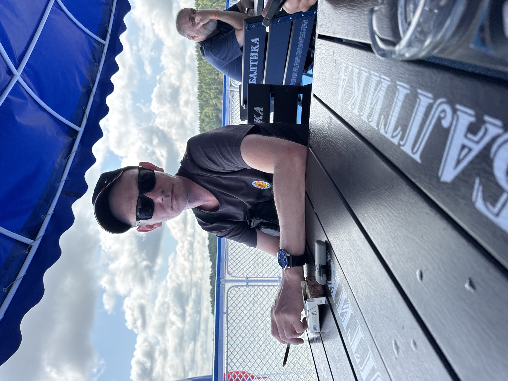 A man wearing a black baseball cap and sunglasses sits at a dark wooden table on a boat. He is holding a cigarette in his left hand and a pack of cigarettes is on the table in front of him, along with a lighter. A watch with a blue face is on his right wrist. Another man with a beard sits at a table behind him. The boat is on a body of water, with a wooded shoreline visible in the distance under a cloudy sky. The text "БАЛТИКА" is visible on the chairs and table. This scene appears to be in Shelanga, Russia.