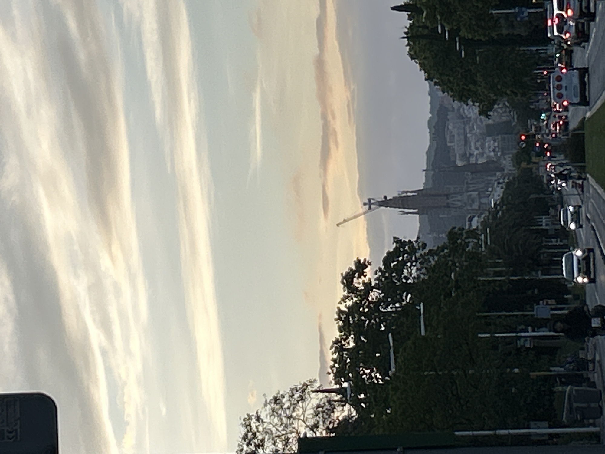 The image depicts a street scene in Barcelona, Spain, with the Sagrada Familia basilica visible in the distance. The sky is overcast with streaky clouds. A construction crane is positioned near the Sagrada Familia. The street is lined with trees and lampposts. There is a road with cars and other vehicles traveling along it. A pedestrian is walking on the sidewalk.
