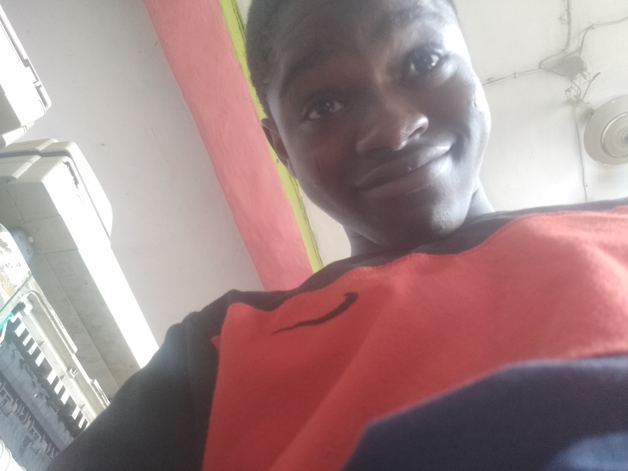 A low-angle selfie of a young Black man with dark skin and short hair. He is smiling slightly. He is wearing a red and black shirt with a dark blue portion visible at the bottom. The photo appears to be taken indoors. Behind the man, there is a wall with a pink stripe and a portion of the ceiling with a ceiling fan. To the left, there are several white electrical boxes or fixtures. The specific location in Shangisha, Nigeria is not discernible from the image.