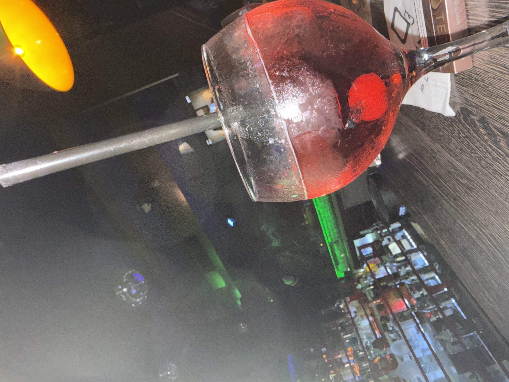 The image shows a partially consumed red cocktail in a large glass with a straw, resting on a dark wooden surface. A red cherry is visible inside the glass. Nearby are boxes and a white napkin. The background shows the interior of a bar or restaurant with a dimly lit ambiance. A distant view of bottles on shelves, illuminated by green and blue lights, can be seen. While the general setting is a bar or restaurant, there is insufficient information to determine if this image was taken in Ruse, Bulgaria. No people or specific actions are directly observable.