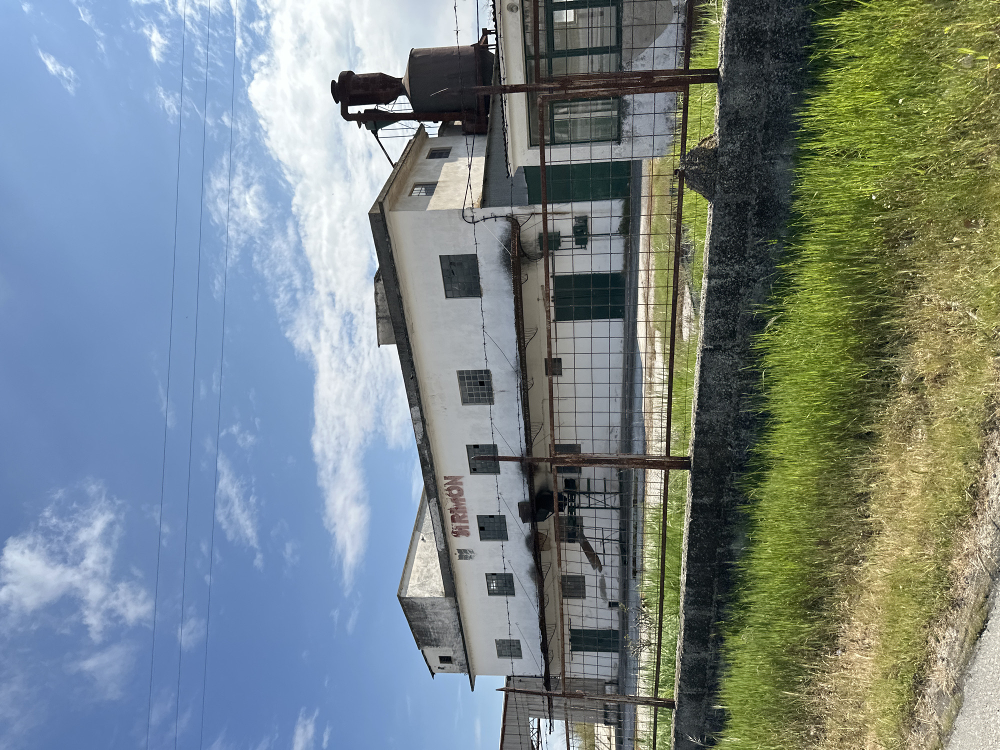 The image shows an old, multi-story, white building, possibly a former industrial site or factory. The building has multiple windows and doors, some of which are painted green. There is some rusty machinery on the roof. The building is surrounded by a rusty wire fence and some vegetation. The sky is blue with scattered clouds. The writing on the building spells out "SIRIMON." Based on the provided location, this is likely the former Sirimon Textile Factory in Lefkonas, Greece.